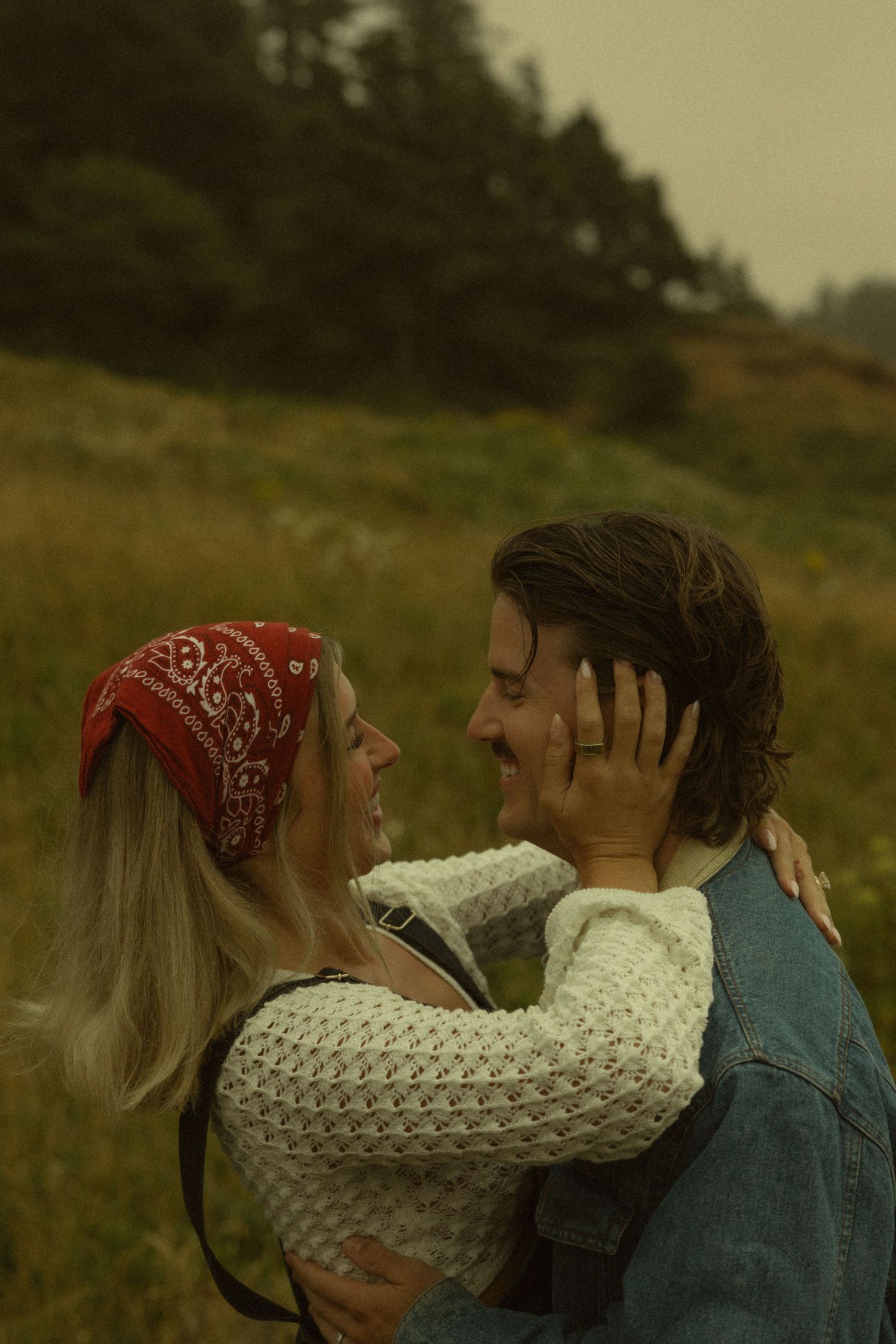 A couple embraces and smiles at each other in a meadow, woman wearing red bandana and white knit sweater.