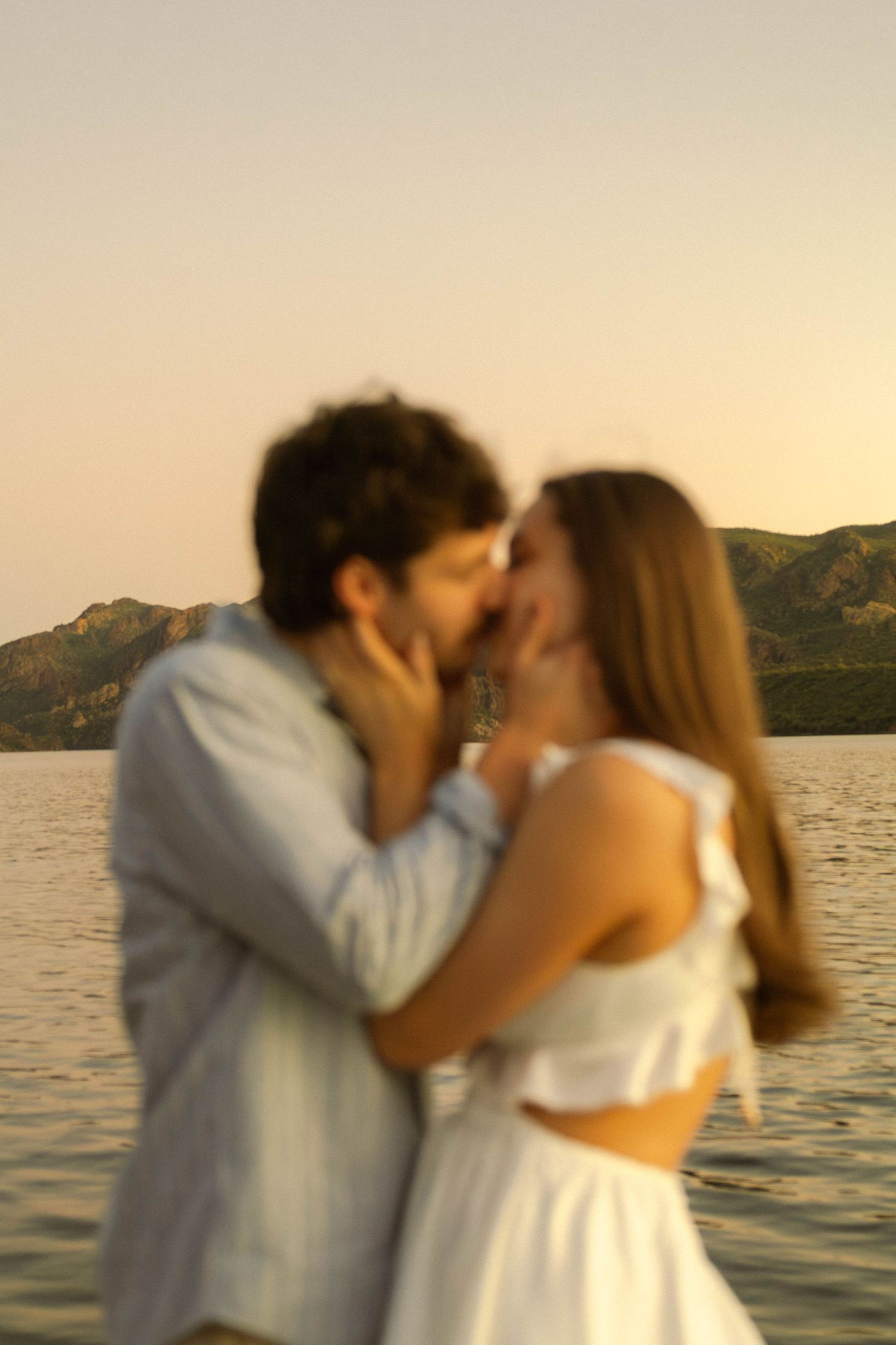 Romantic couple sharing an intimate kiss on a dock at golden hour with mountains behind them.
