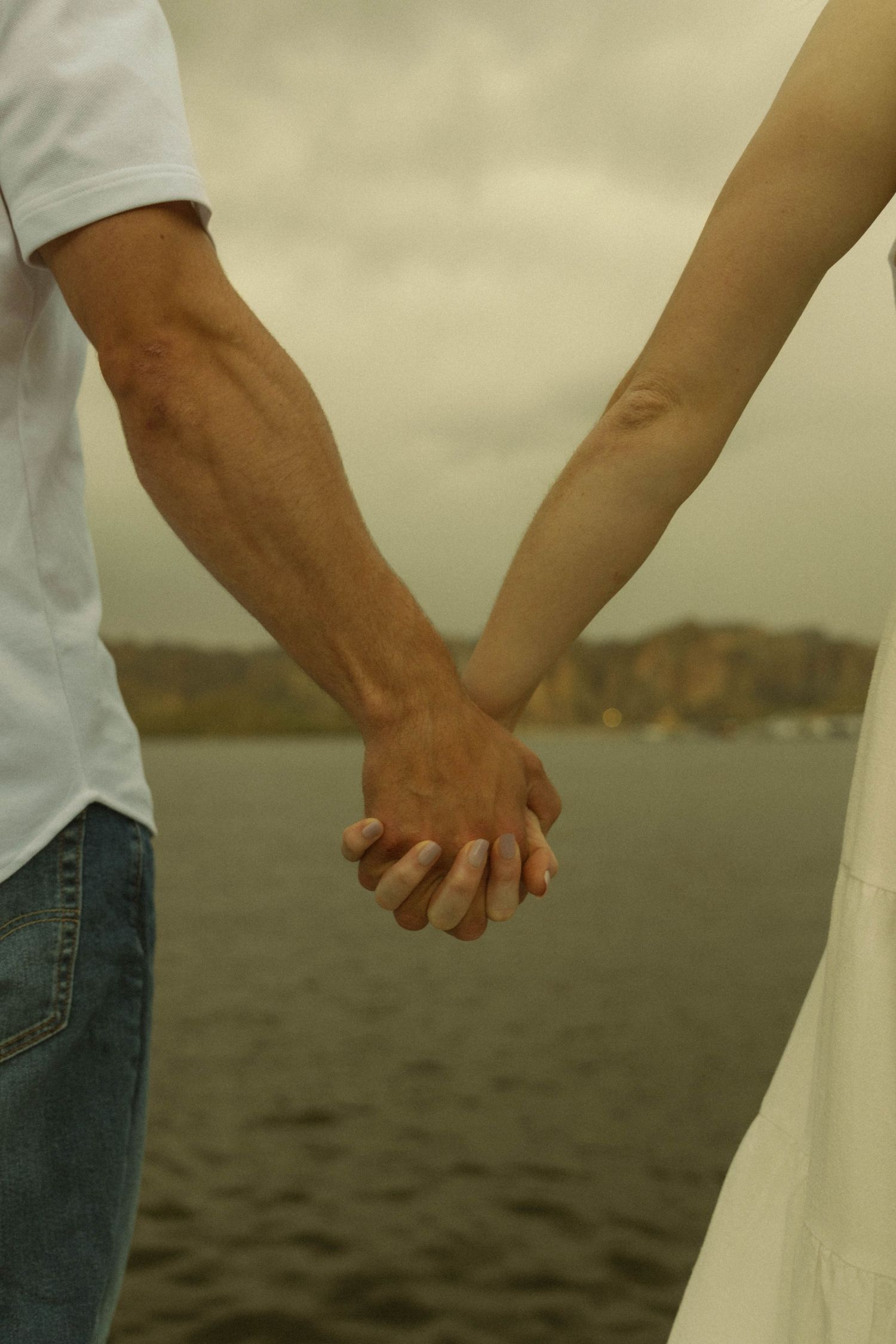 A detailed shot of two people holding hands against a waterfront mountain landscape.