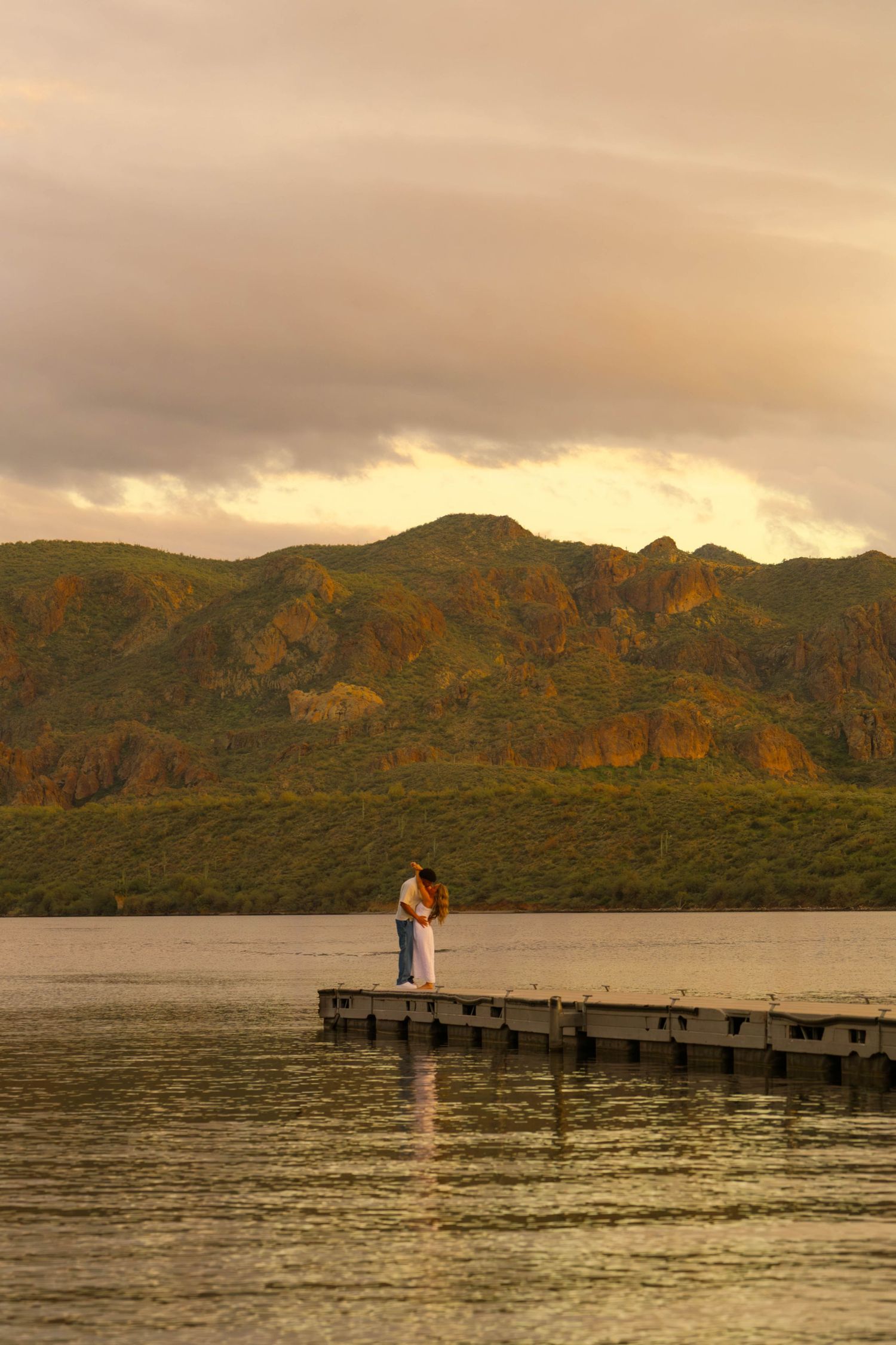 A couple stands on a wooden pier at sunset with mountains in the background creating a romantic silhouette.