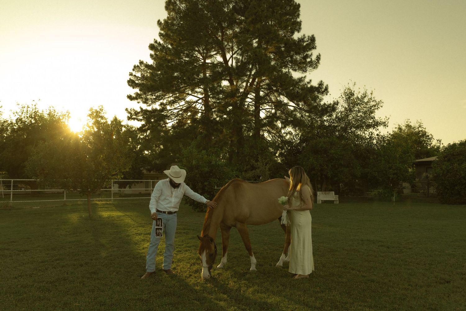 A cowboy and woman in a white dress stand with a tan horse on a green lawn at golden hour.