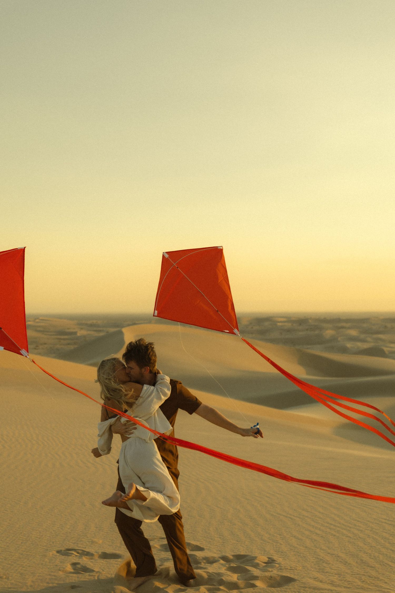 Couple embracing on golden sand dunes at sunset with two red kites soaring above in a romantic desert scene.