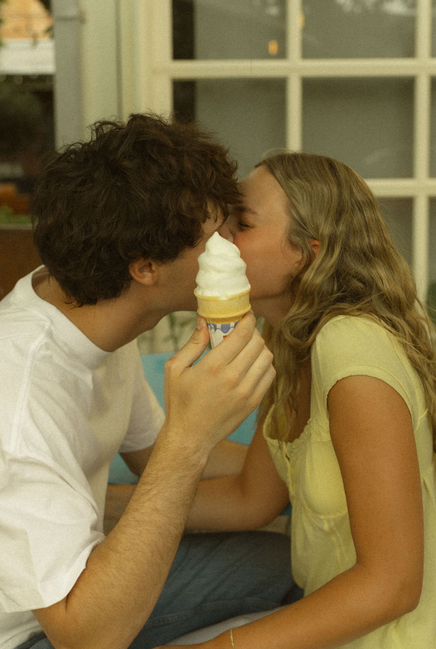 Young couple sharing an ice cream cone kiss outdoors on a sunny day.