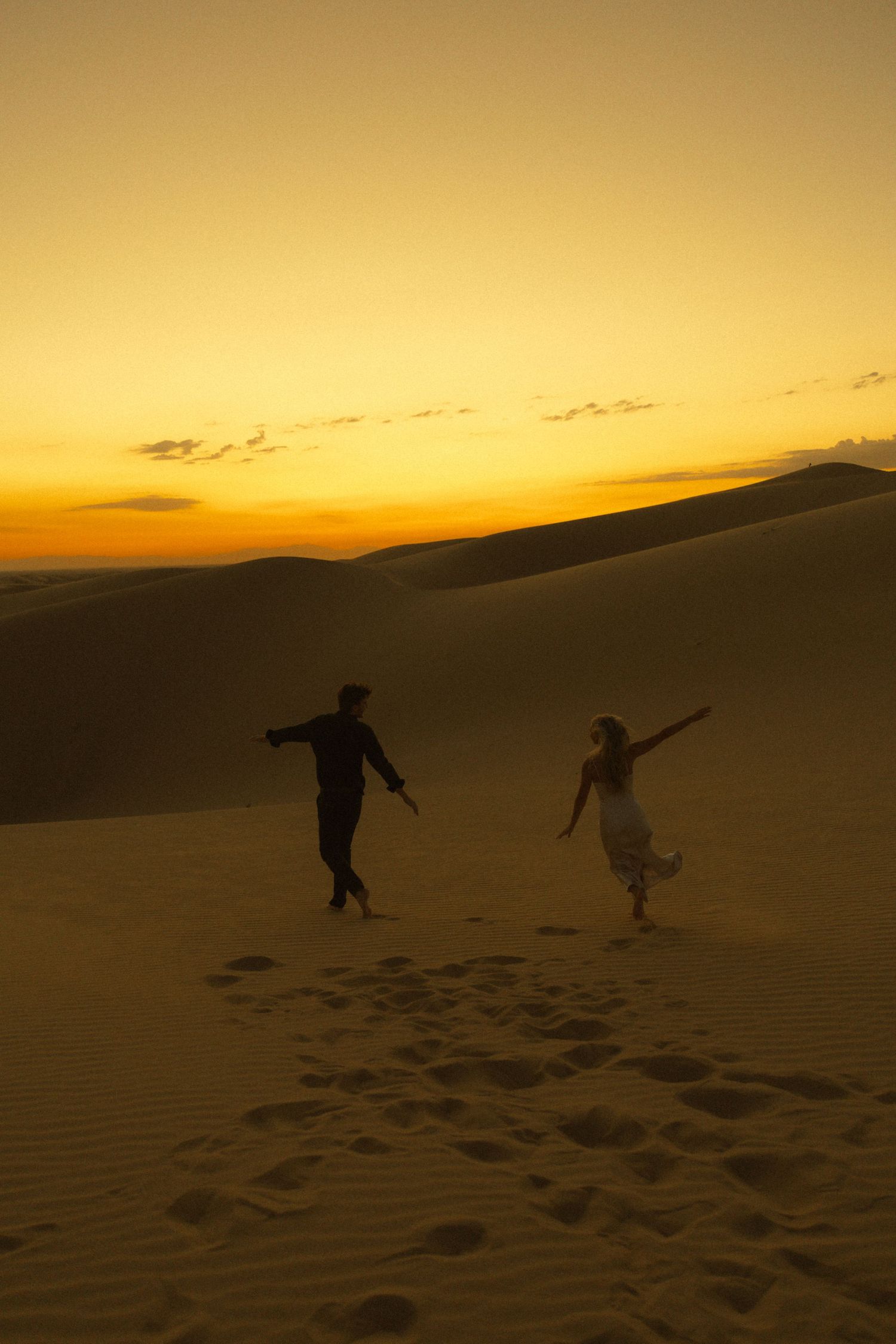 Silhouette of couple dancing freely on sand dunes against a vibrant orange desert sunset sky.