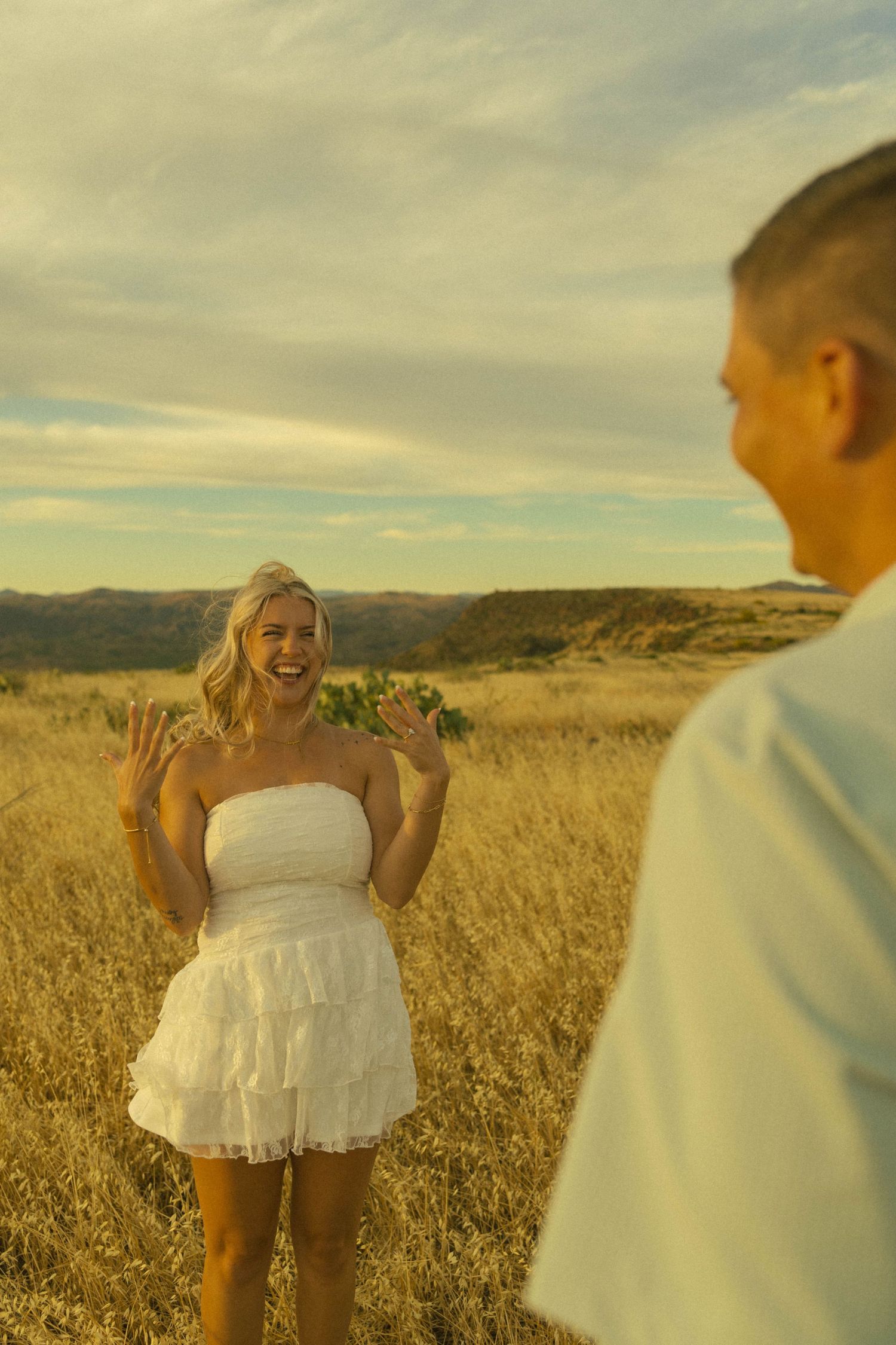Joyful blonde woman in white dress laughing with hands raised in sunny field.