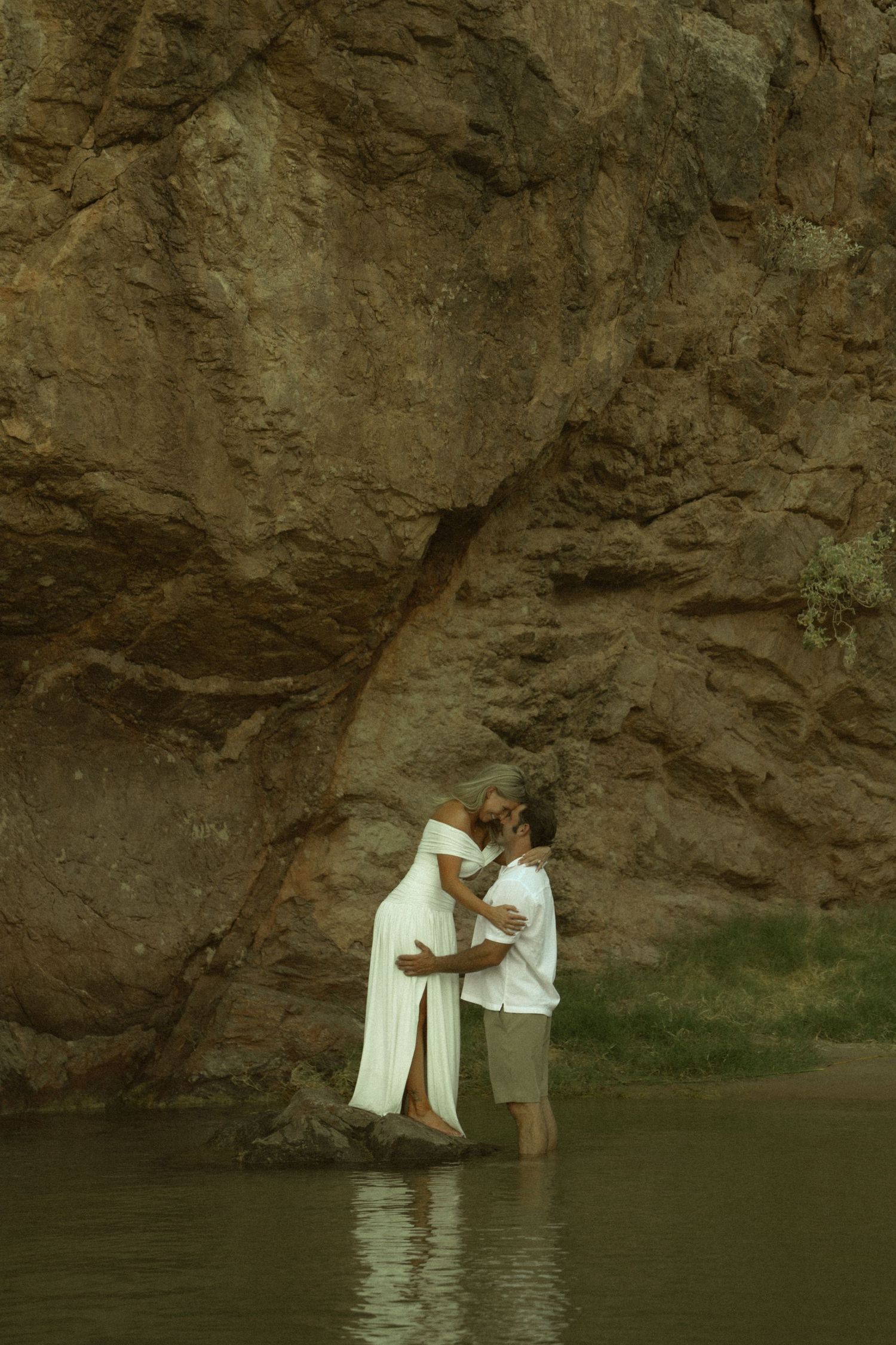 Couple sharing a romantic kiss while standing in a river with a dramatic rocky cliff backdrop.