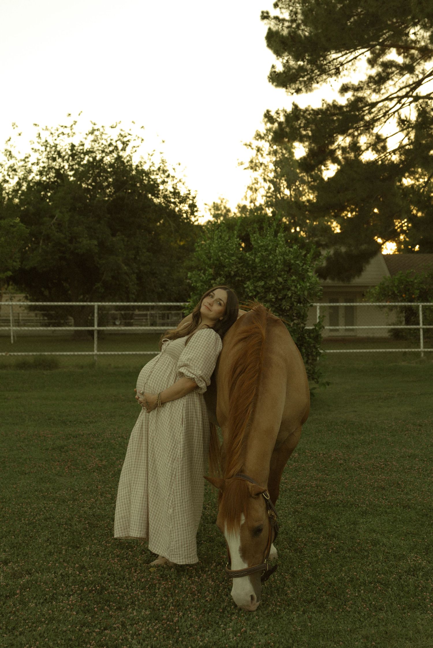 Pregnant woman in white dress leaning against a grazing horse at sunset in a lush green pasture.