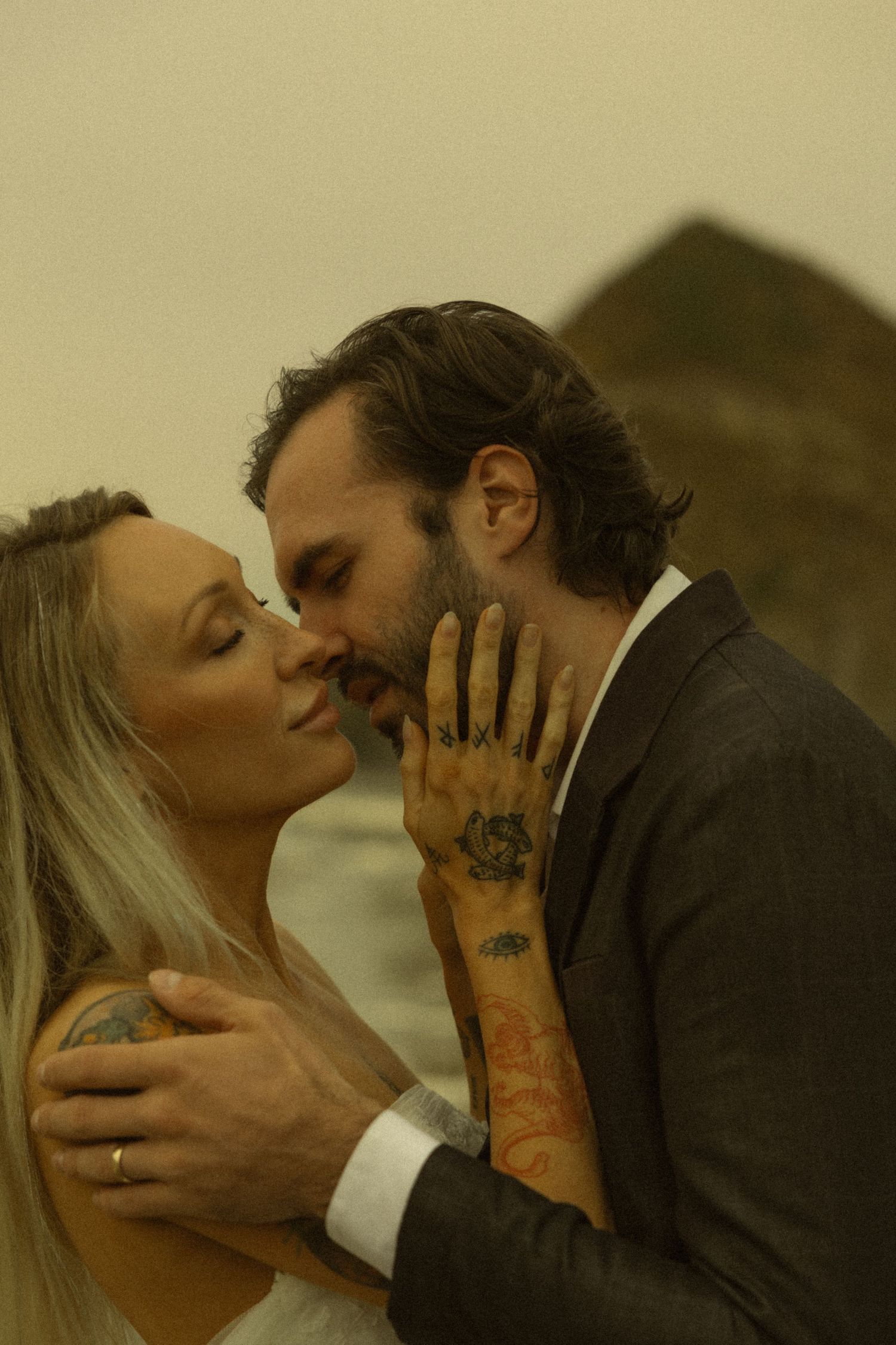 A couple shares a romantic kiss, the groom in a suit with tattooed hands embracing a blonde bride outdoors.