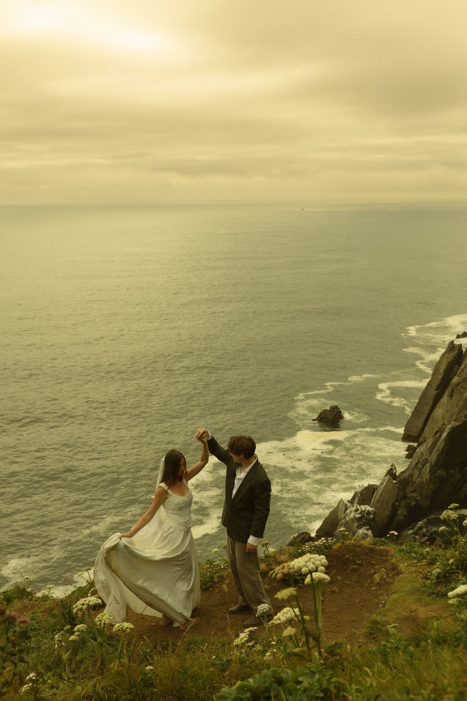 Couple in wedding attire dancing on a coastal cliff overlooking the ocean at sunset.