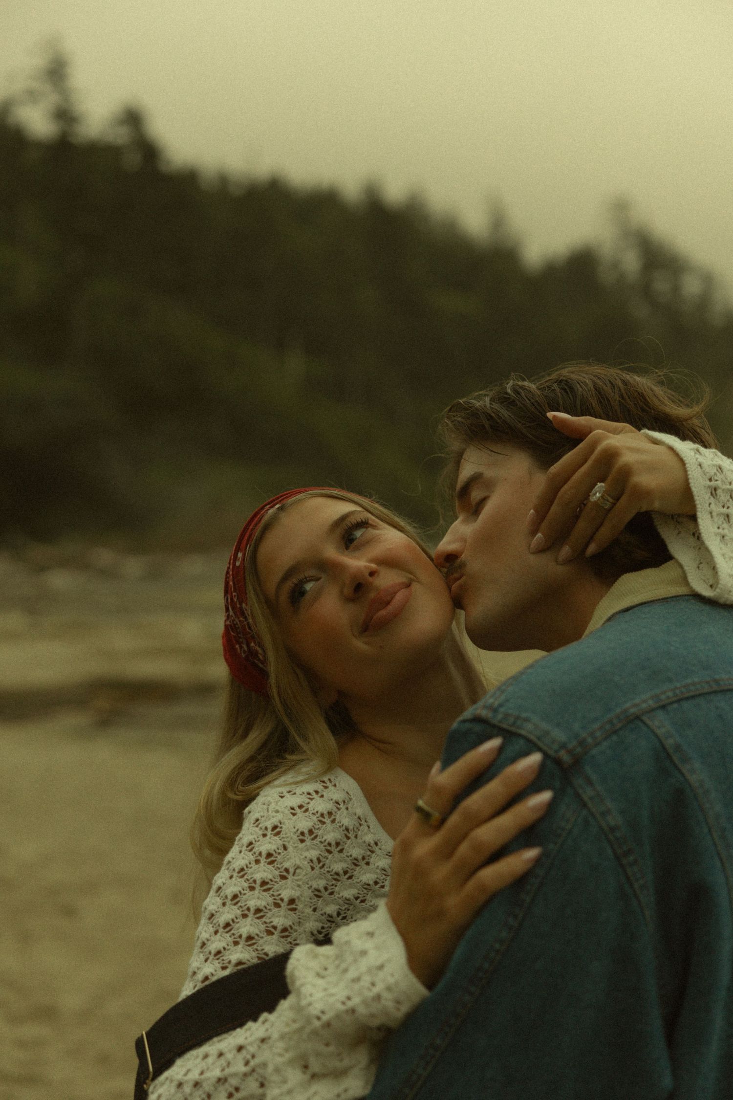 A man kisses a smiling woman on the cheek on a foggy beach, she wears a red bandana and white sweater.