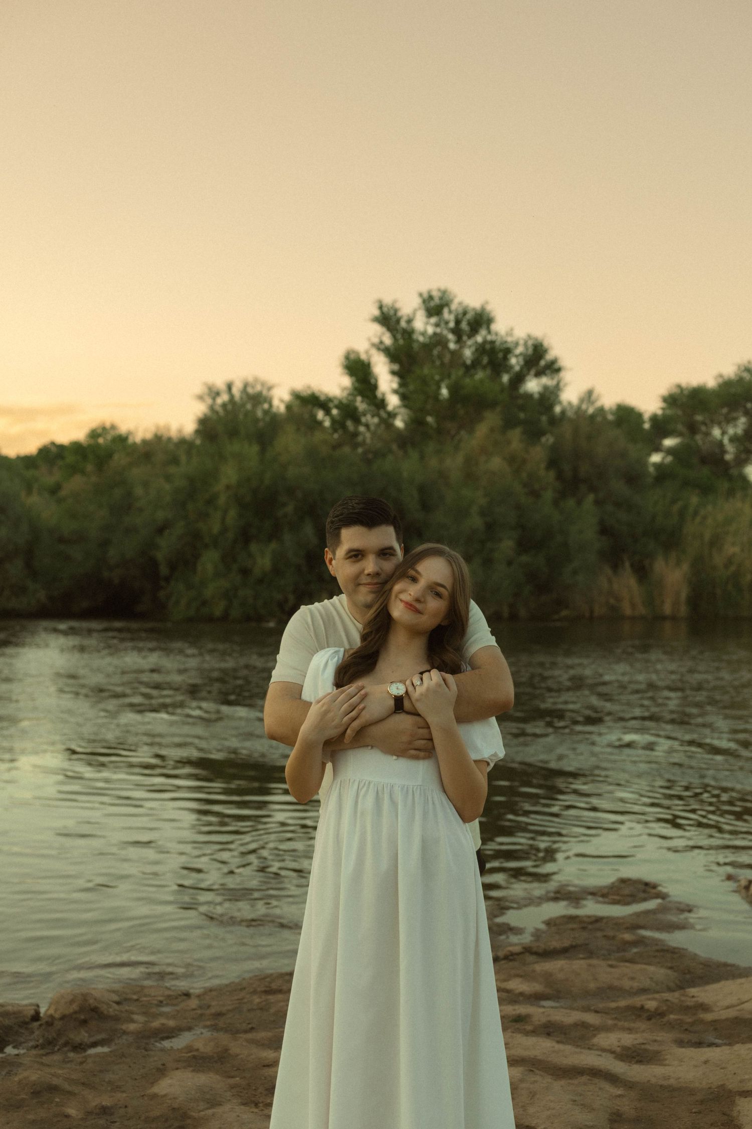 Couple embracing at sunset by a river, woman in white dress, man in white shirt, with lush green trees in background.
