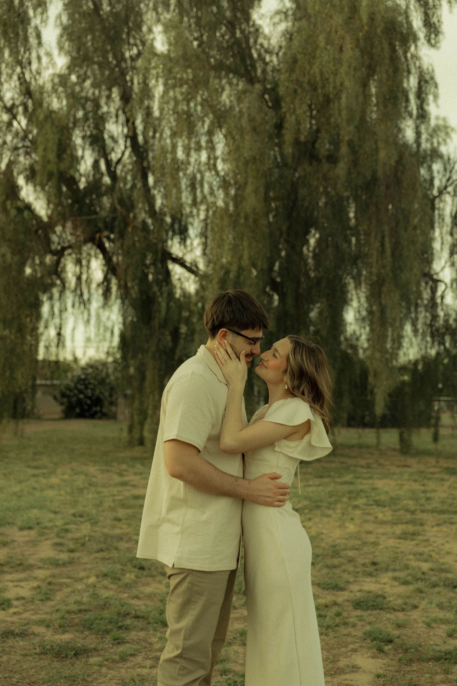 Couple embracing and almost kissing while standing under a large weeping willow tree.