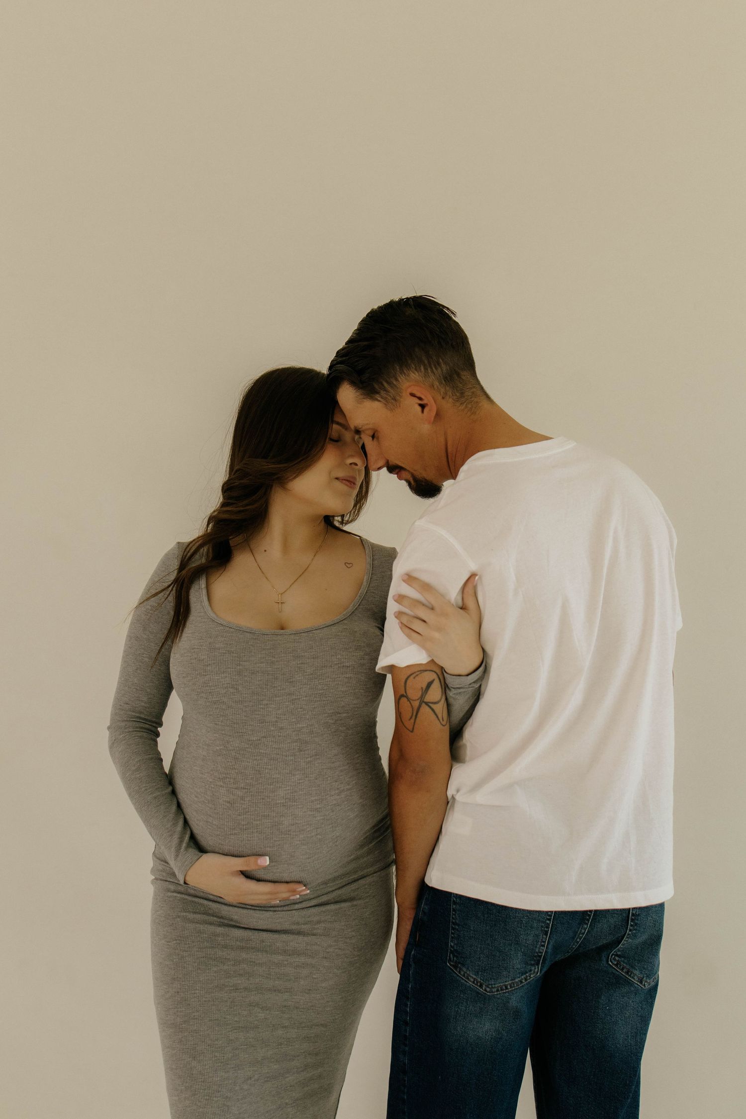 An expectant couple in casual clothing embraces tenderly against a neutral wall during a maternity photoshoot.