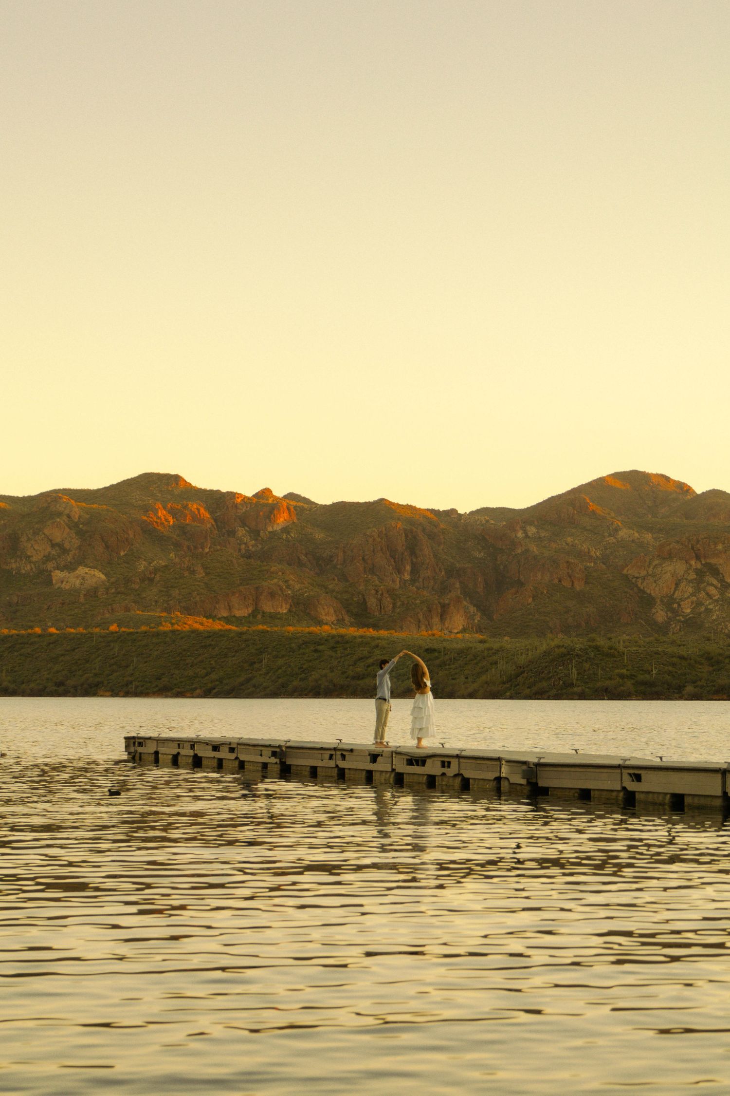 Two figures standing on a wooden pier during golden hour with mountains in the background.