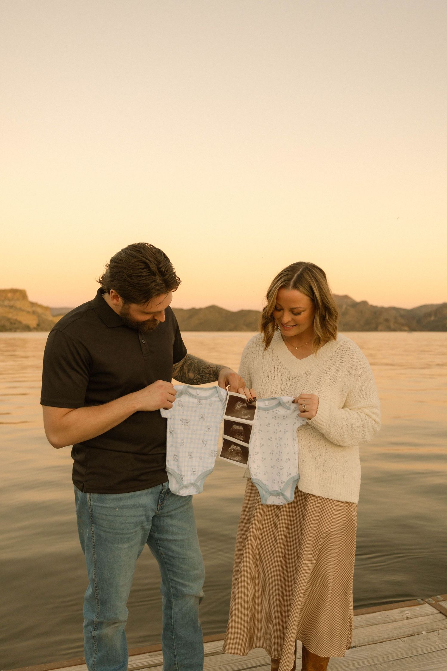 Expectant couple on dock holding baby clothes and sonogram pictures at sunset.