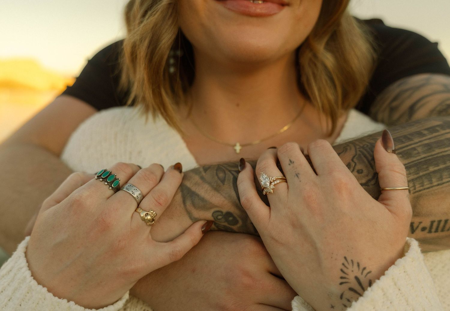 Close-up of hands with multiple rings and a small tattoo resting on an acoustic guitar during golden hour.