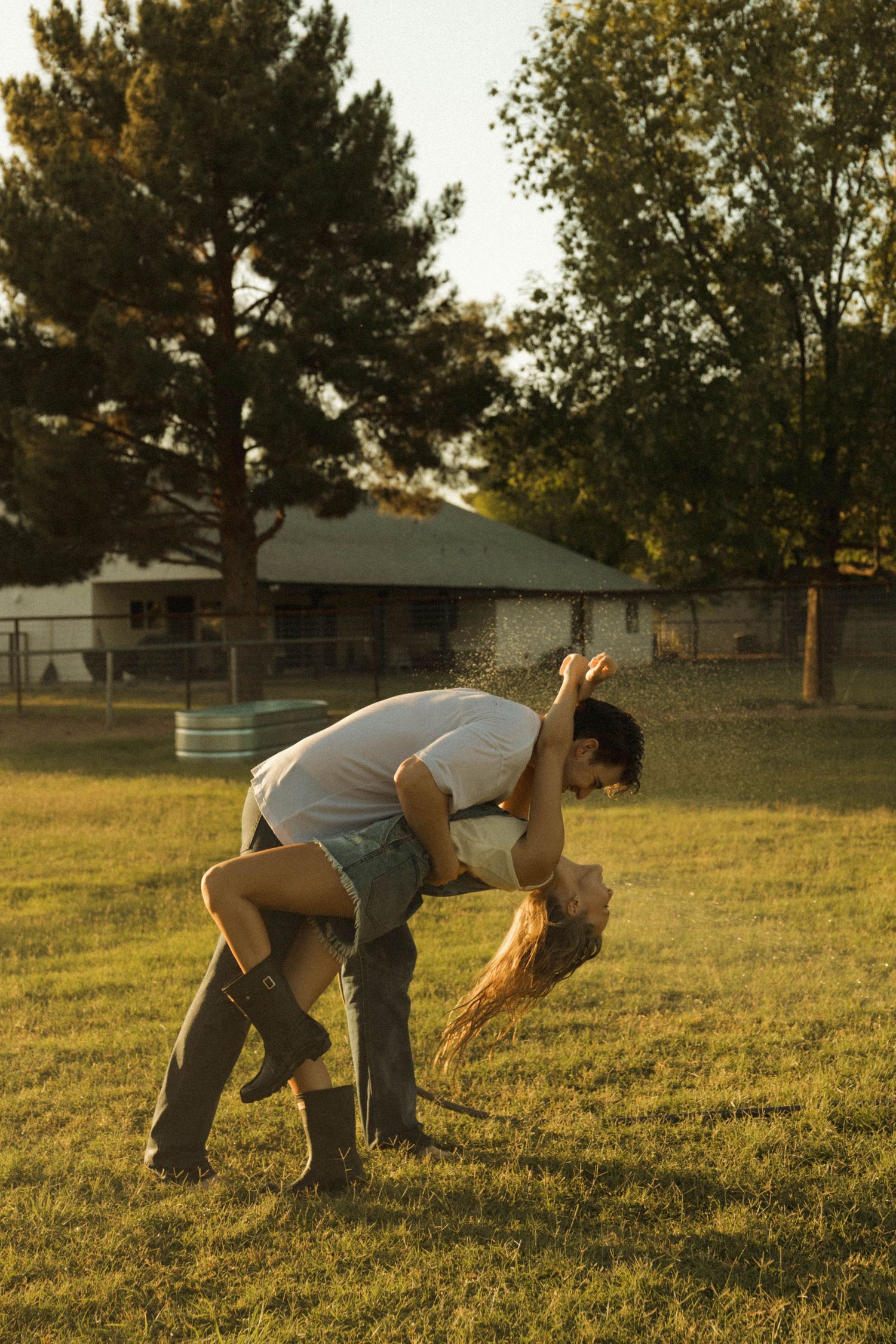 Couple sharing a passionate dip kiss on a green lawn with water spray in golden sunlight.