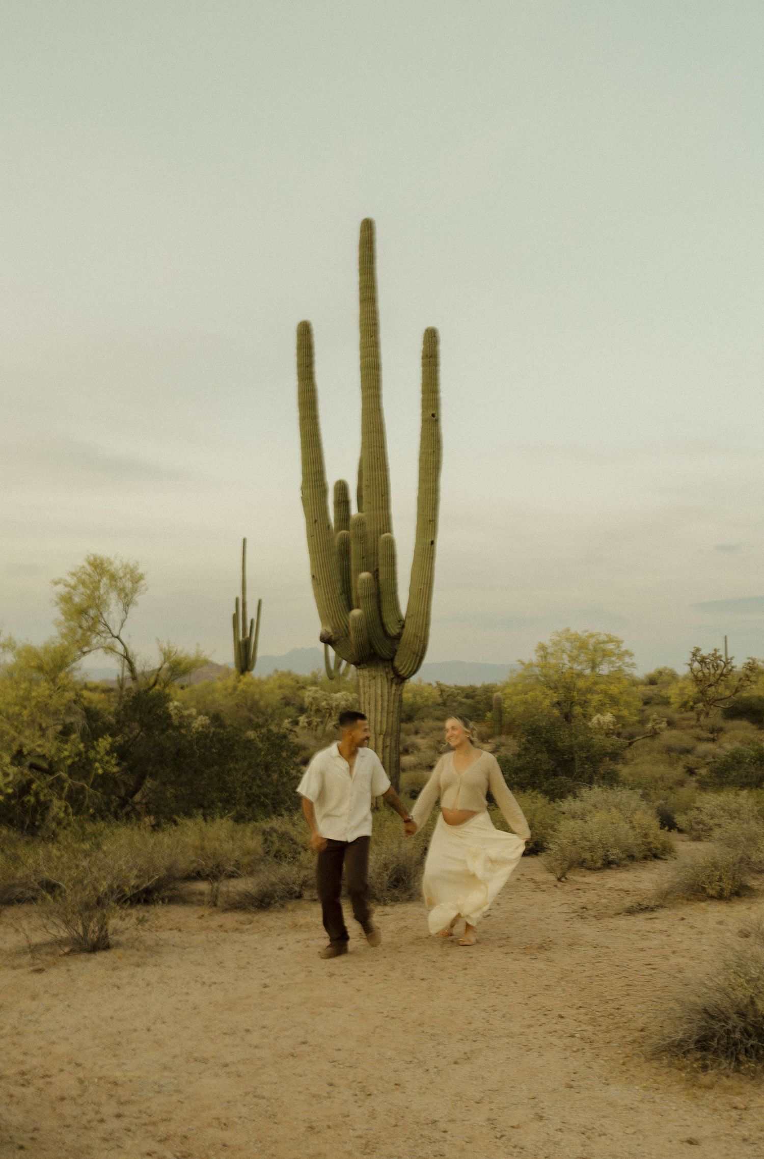 Couple holding hands walking in Sonoran Desert with tall saguaro cactus backdrop at dusk.