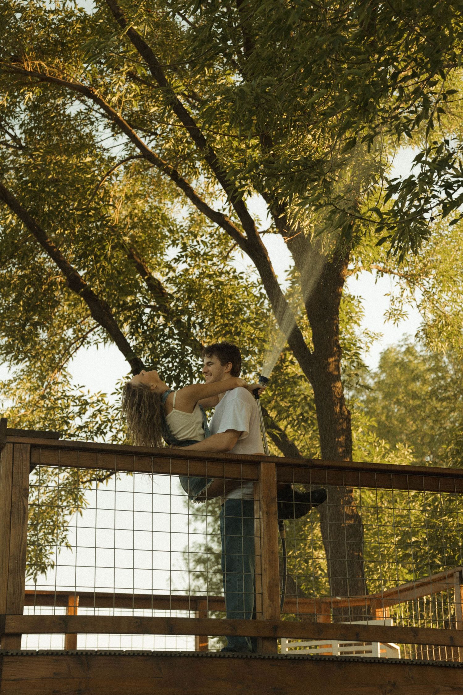 Romantic couple embracing on a wooden deck surrounded by lush green trees in warm sunlight.