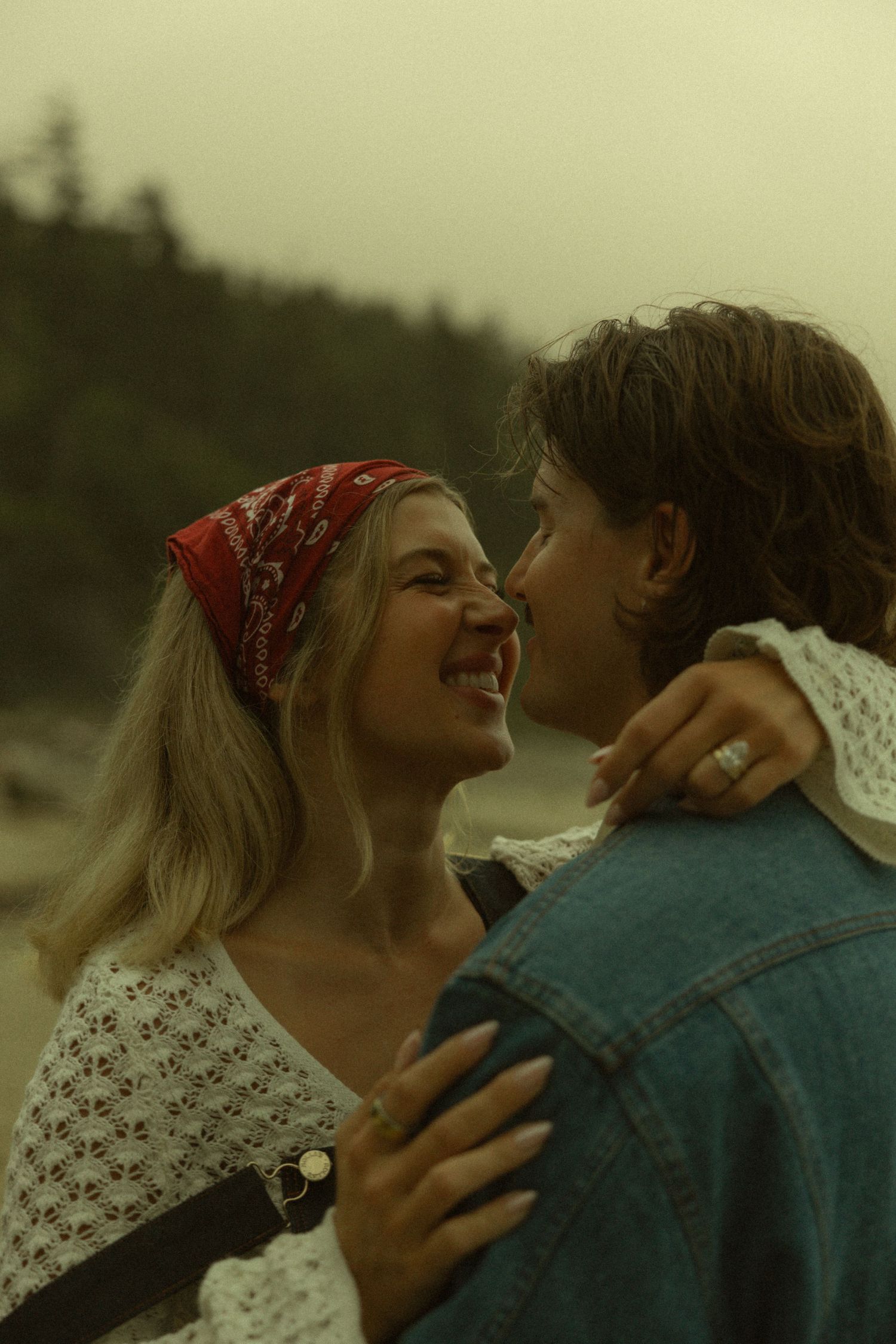 A smiling blonde woman in a red bandana embraces a man in a denim jacket on a misty beach.