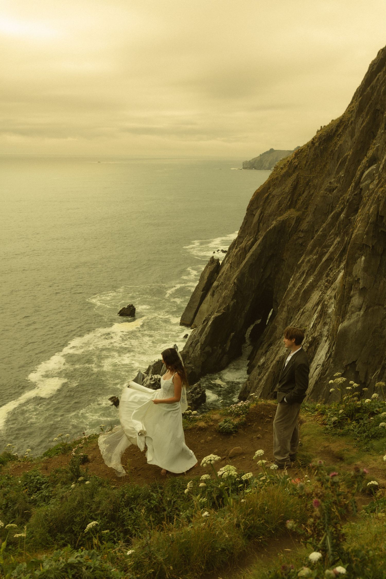 Bride and groom exchange vows on a dramatic coastal cliff overlooking the ocean at sunset.