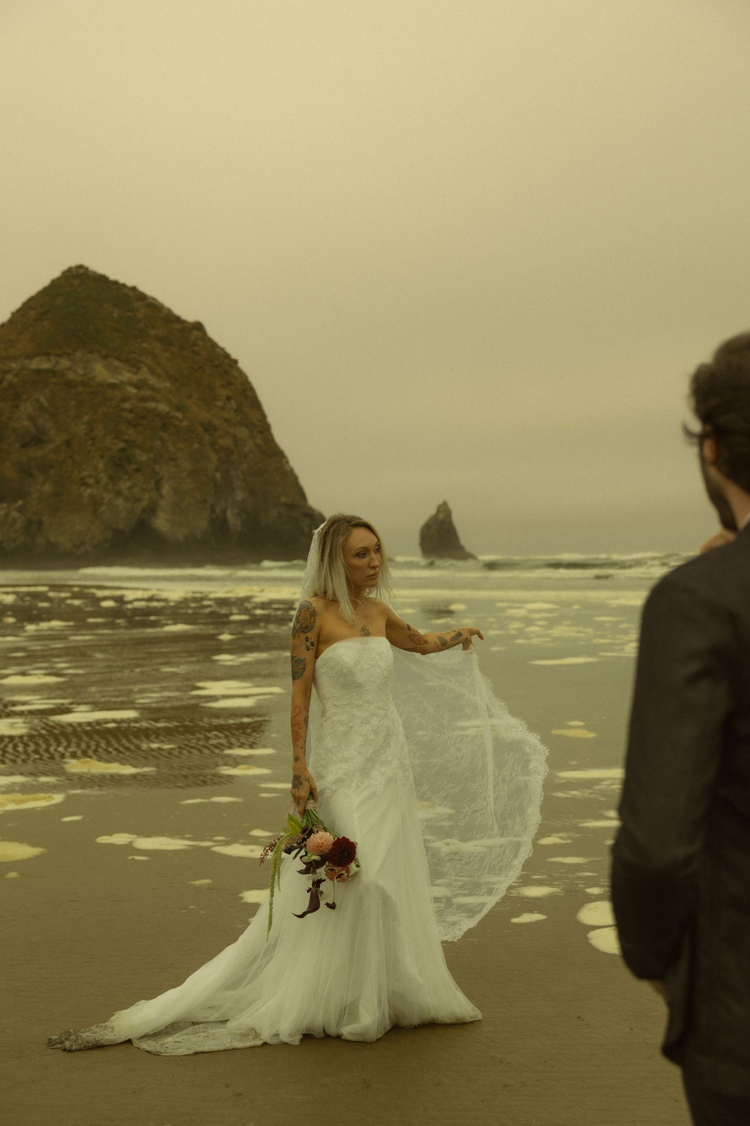 Bride in white gown holds bouquet on misty beach with Haystack Rock, as groom watches in foreground.