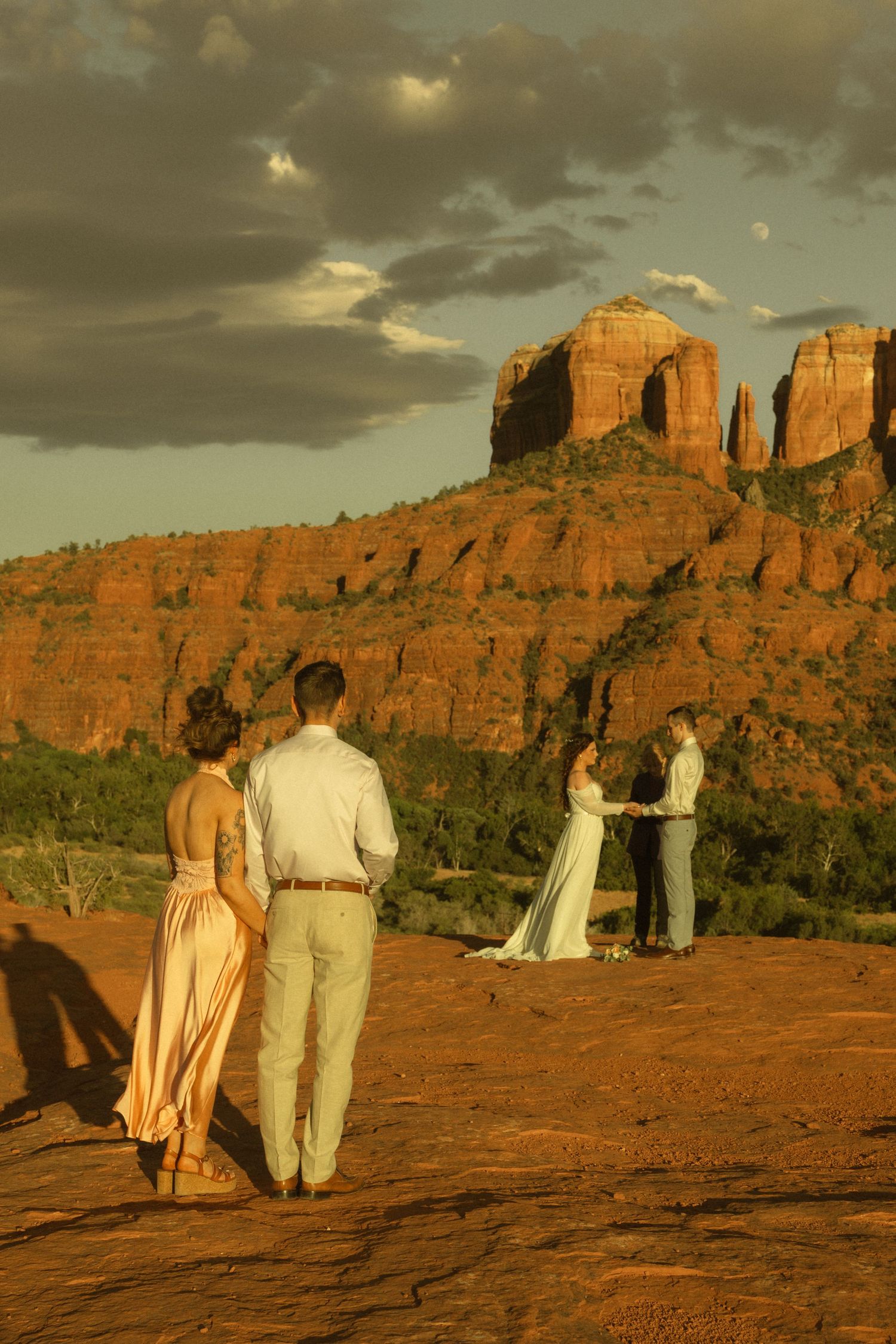 Couple exchanges vows at sunset on red rock formation in Sedona, Arizona, with dramatic buttes and cloudy sky backdrop.
