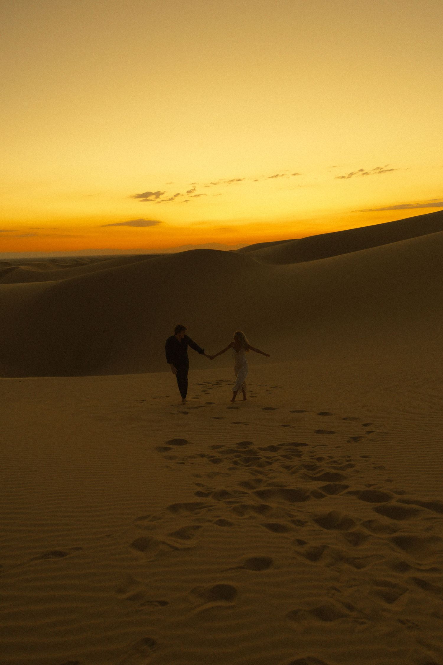 Silhouette of couple holding hands walking on sand dunes at vibrant orange sunset.