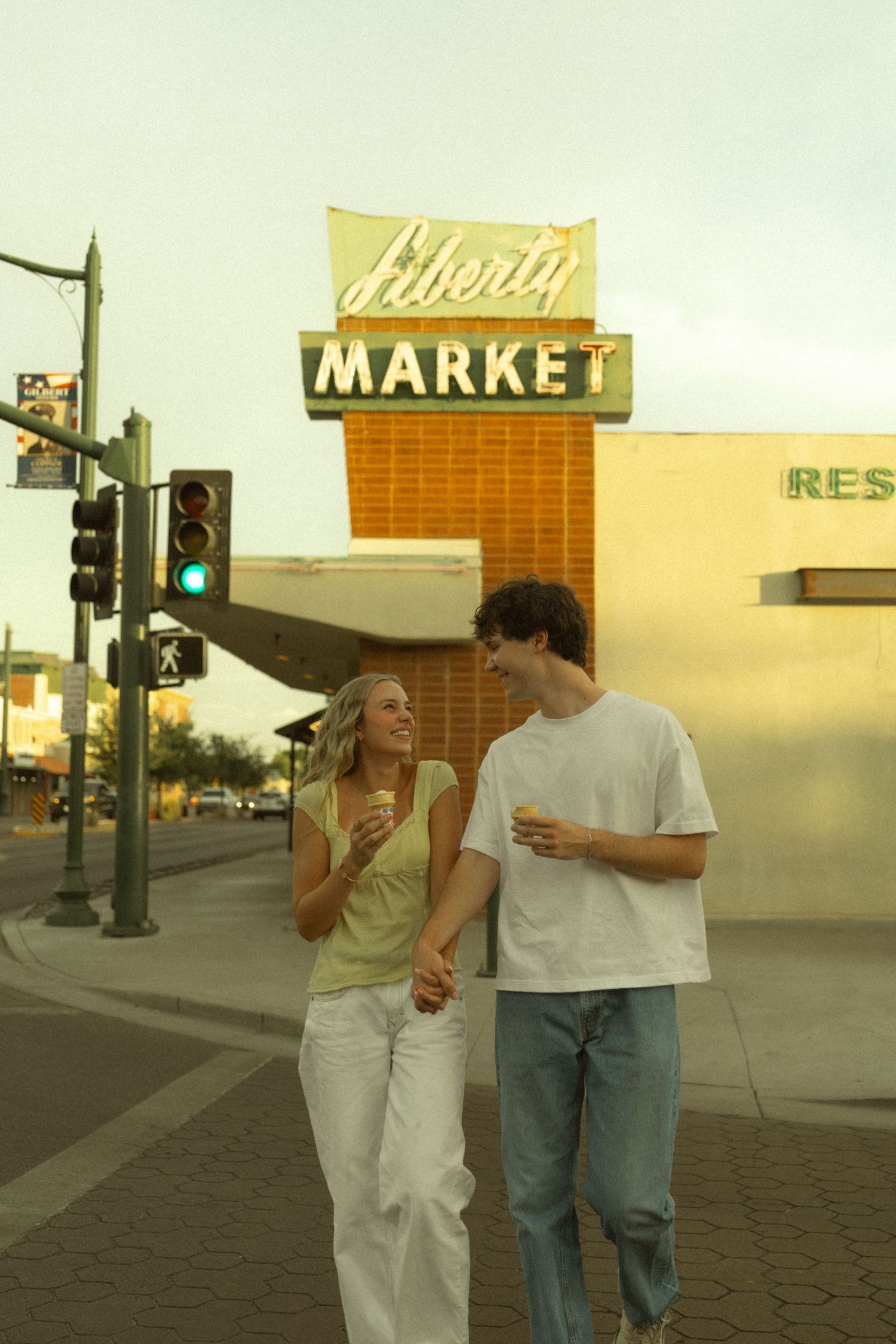 Happy young couple walking hand in hand in front of the Liberty Market sign at golden hour.