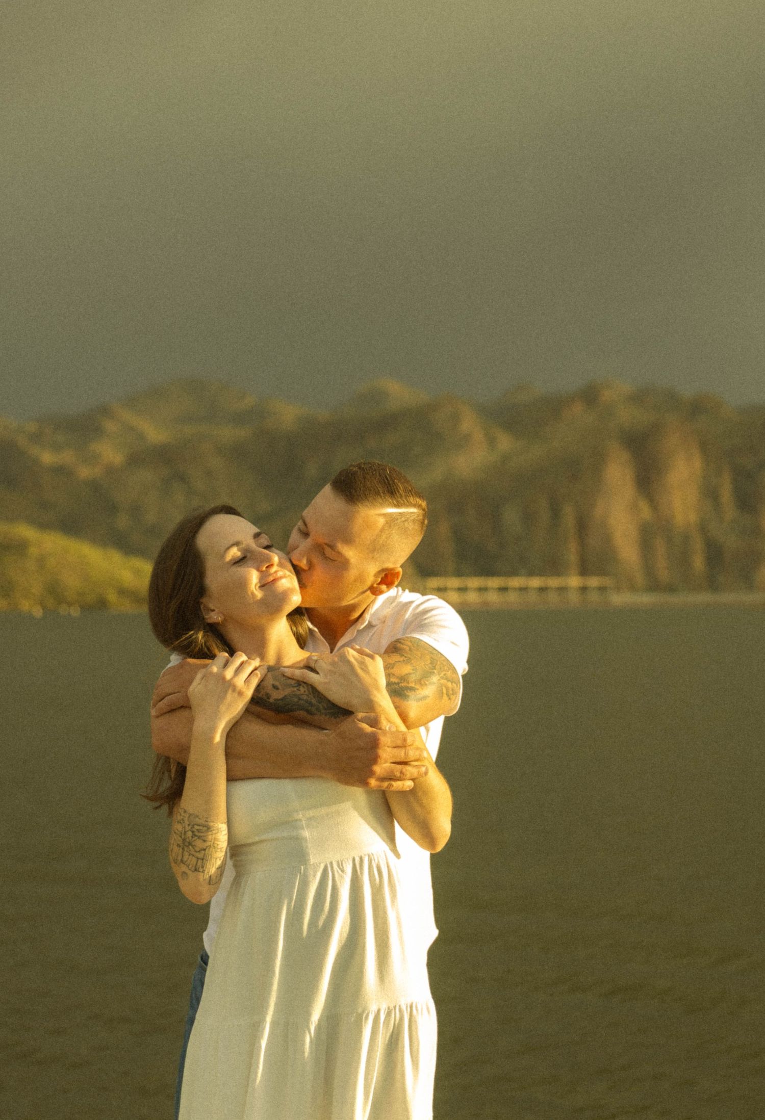 A person in a white dress drinks from a glass bottle against a golden sunset landscape.