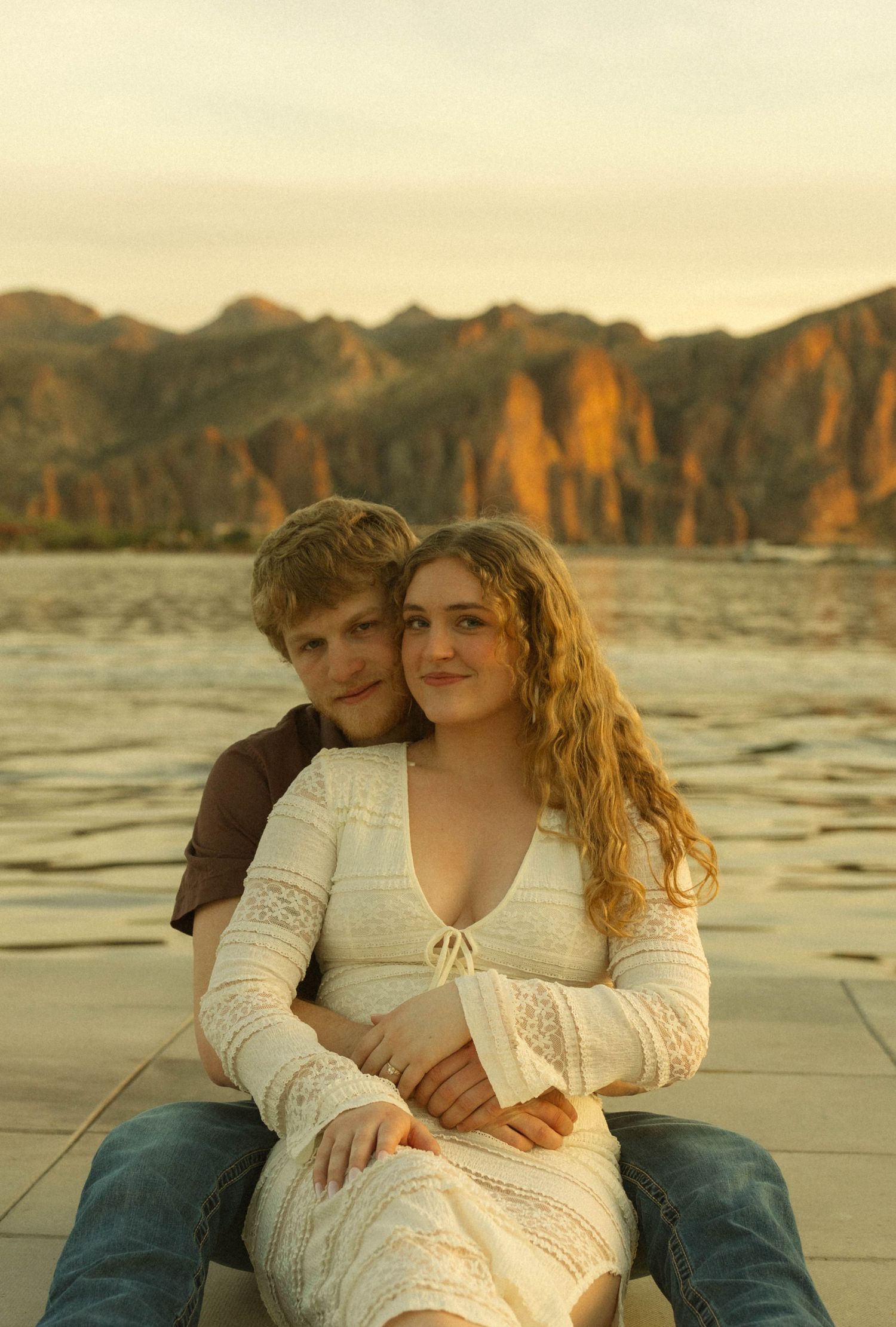 A couple sitting close together on a beach at sunset with mountains visible in the distance.