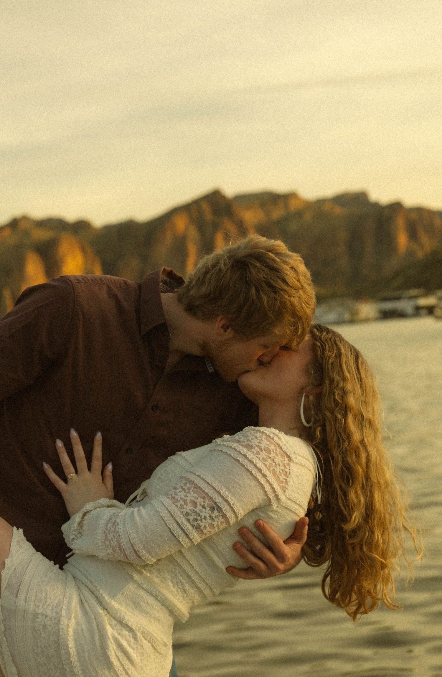 A romantic couple embracing and kissing at sunset by the water with mountains in the background.