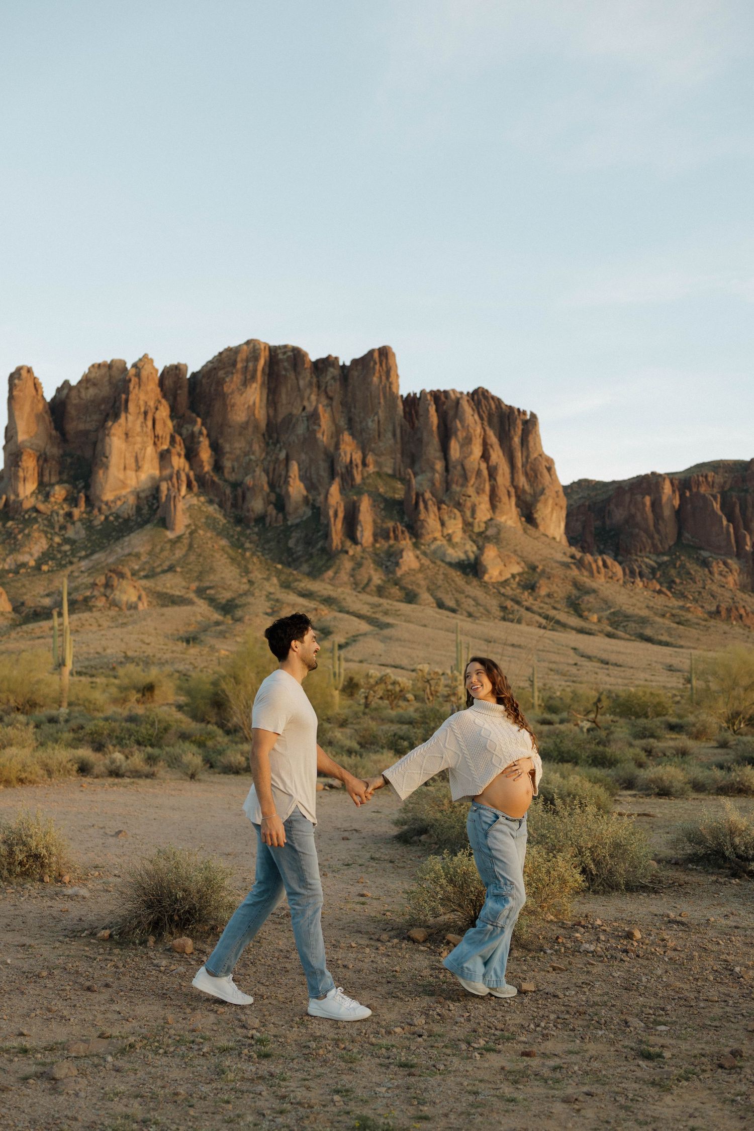 A couple playfully interacts against the dramatic backdrop of rugged desert cliffs and mountains at sunset.