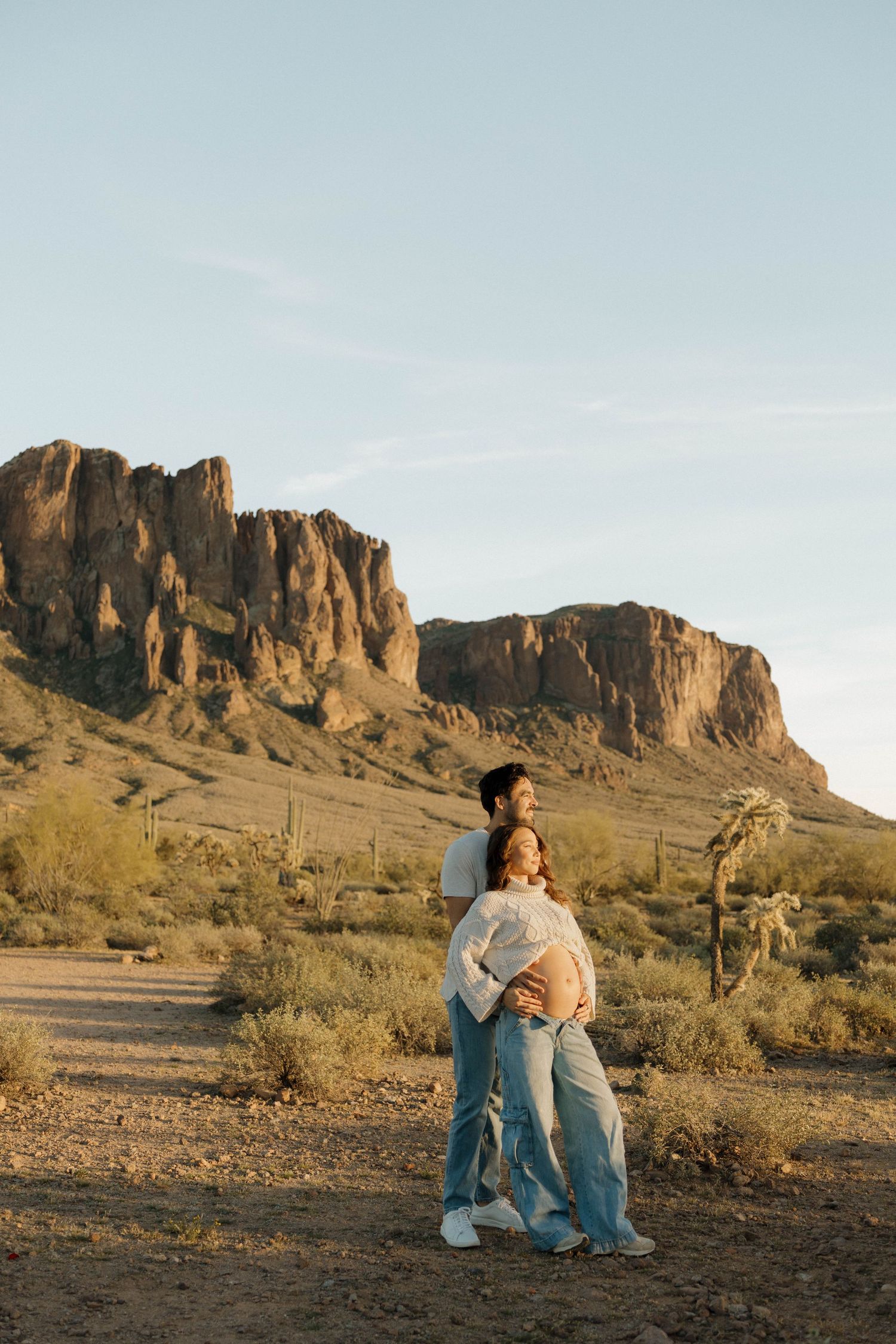A couple sits together against a backdrop of dramatic desert cliffs and mountains at sunset.