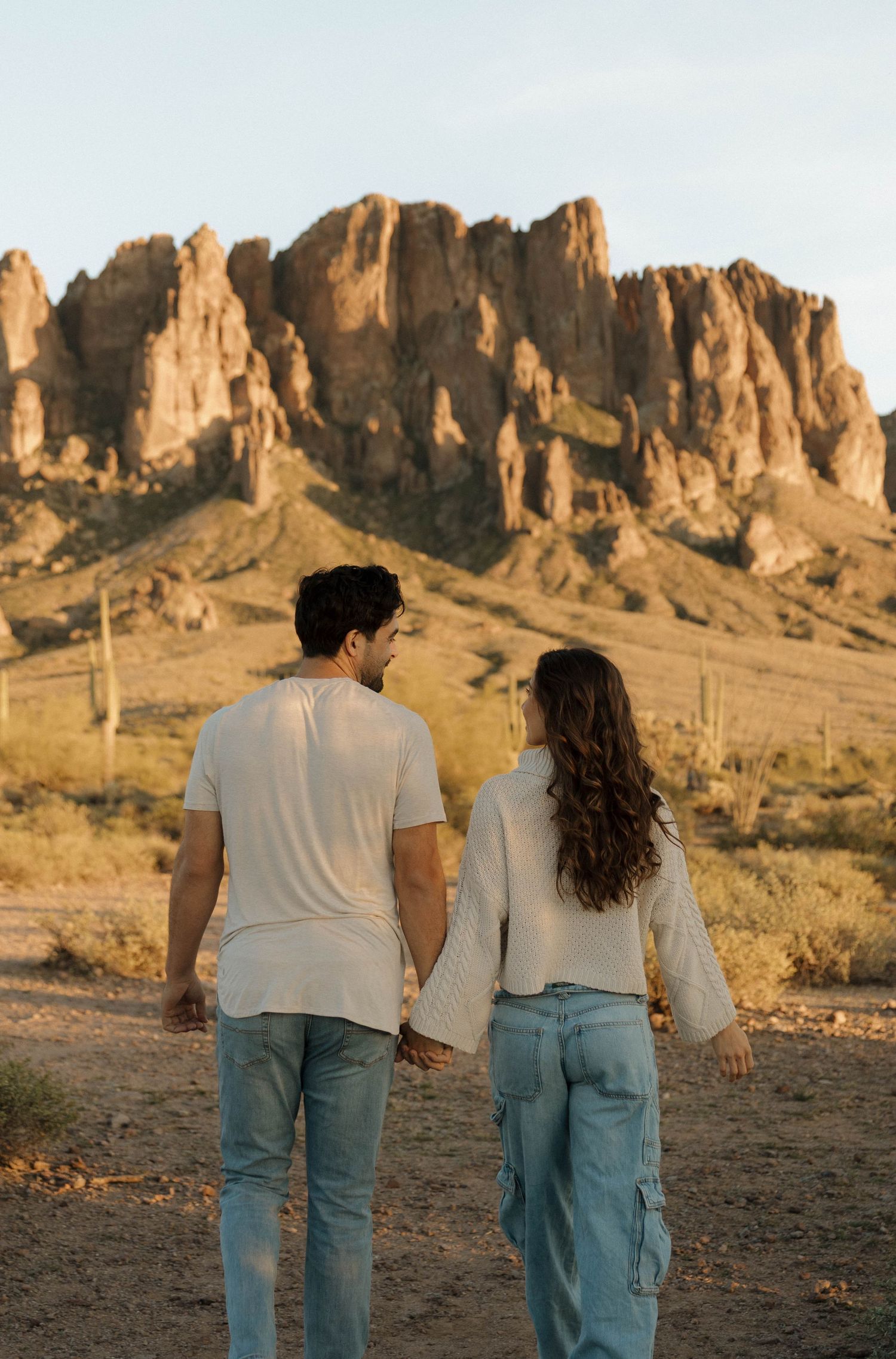 Two people walk together in the desert landscape with towering rock formations of the Superstition Mountains behind them.