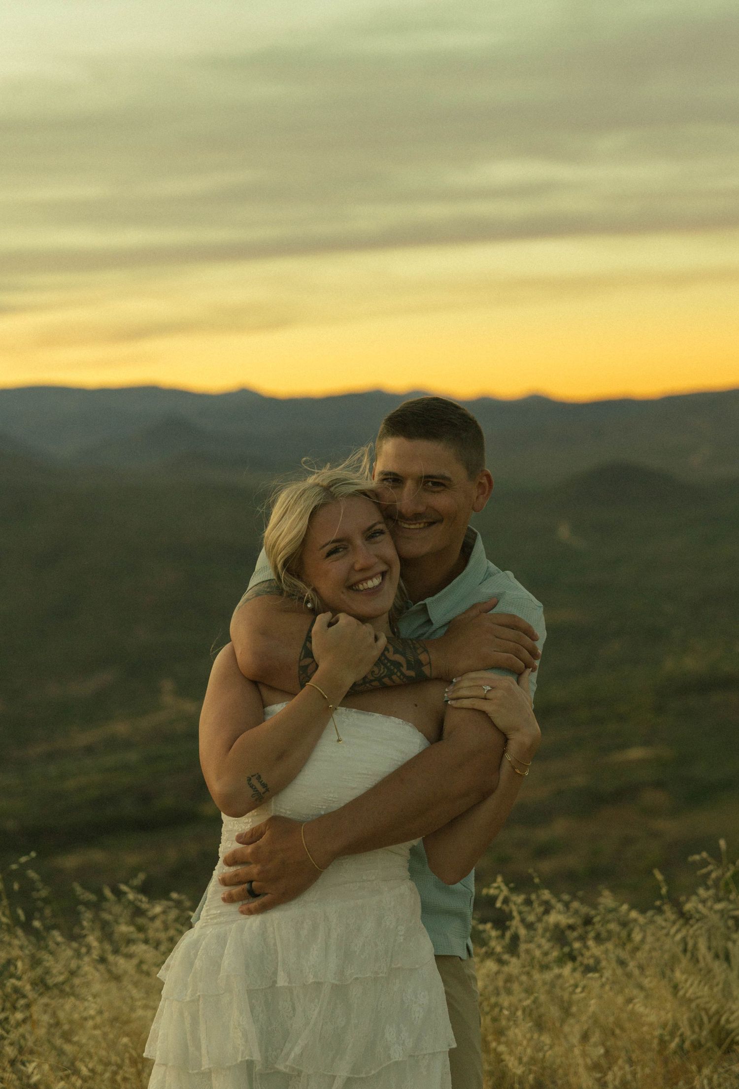 Couple embracing at golden hour sunset with mountain valley views, woman in white dress smiling.