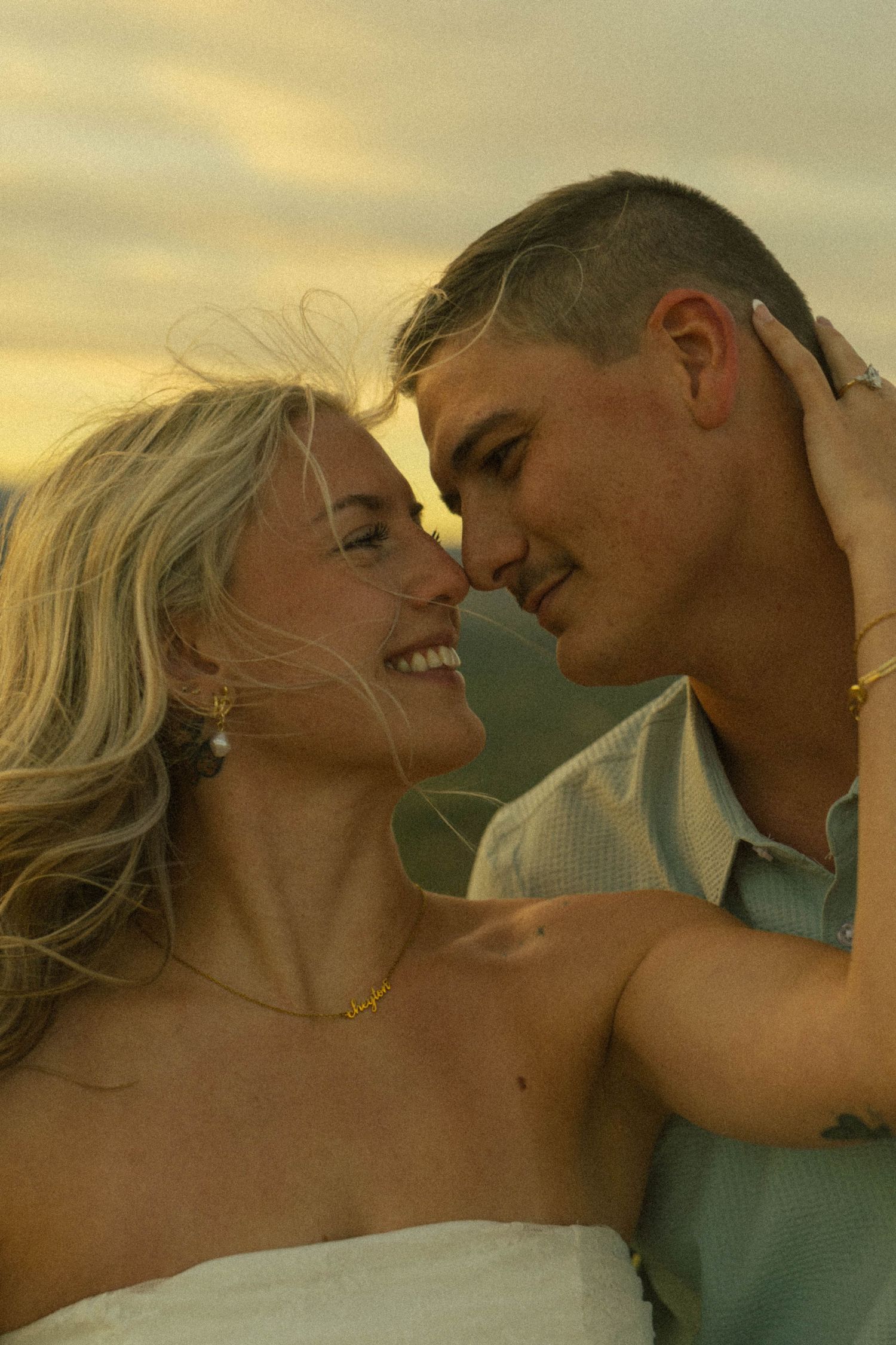 A smiling couple shares a romantic moment at sunset, with the woman in white and the man in a light blue shirt.