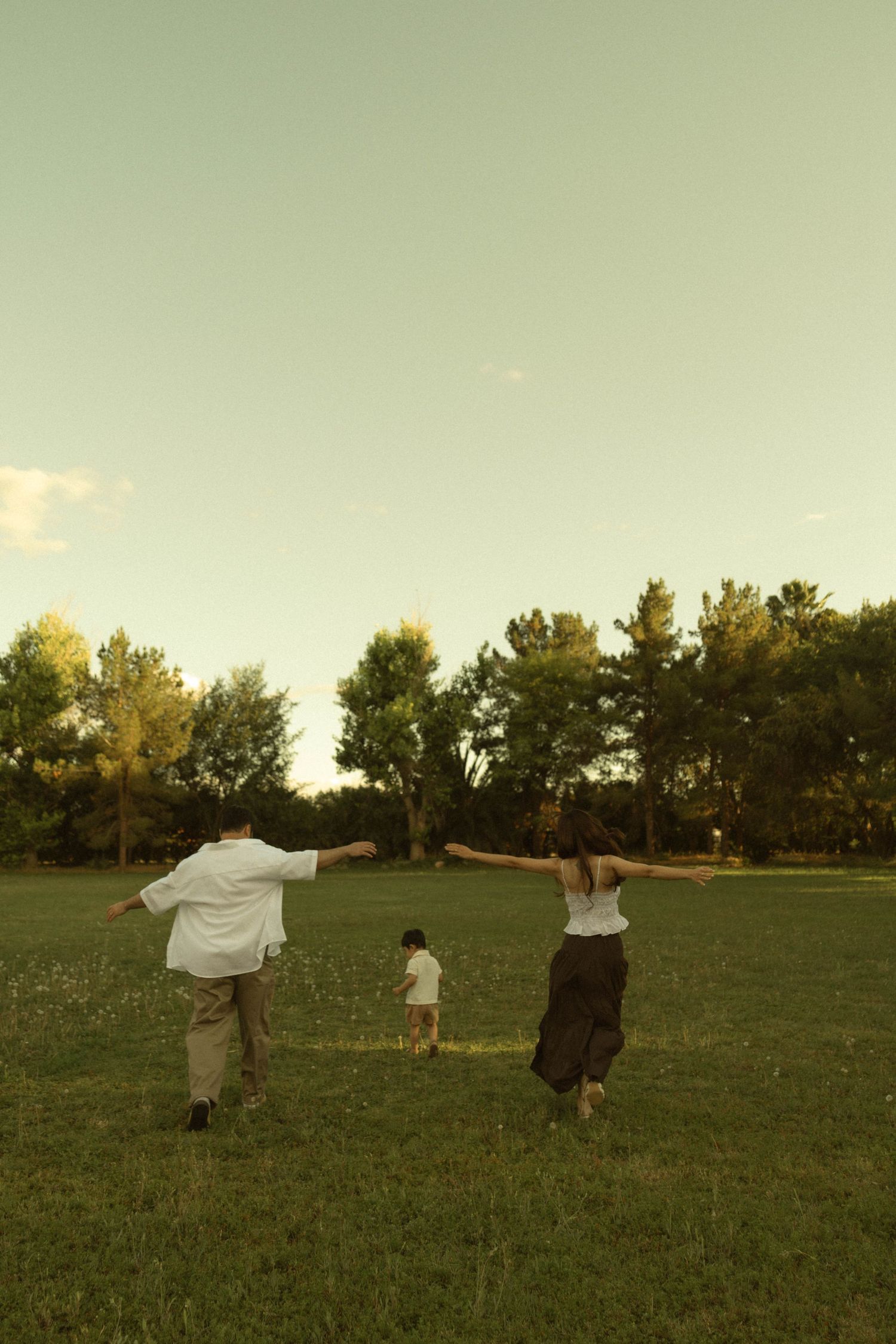 Family of three running joyfully across a green field at sunset, with trees in the background.