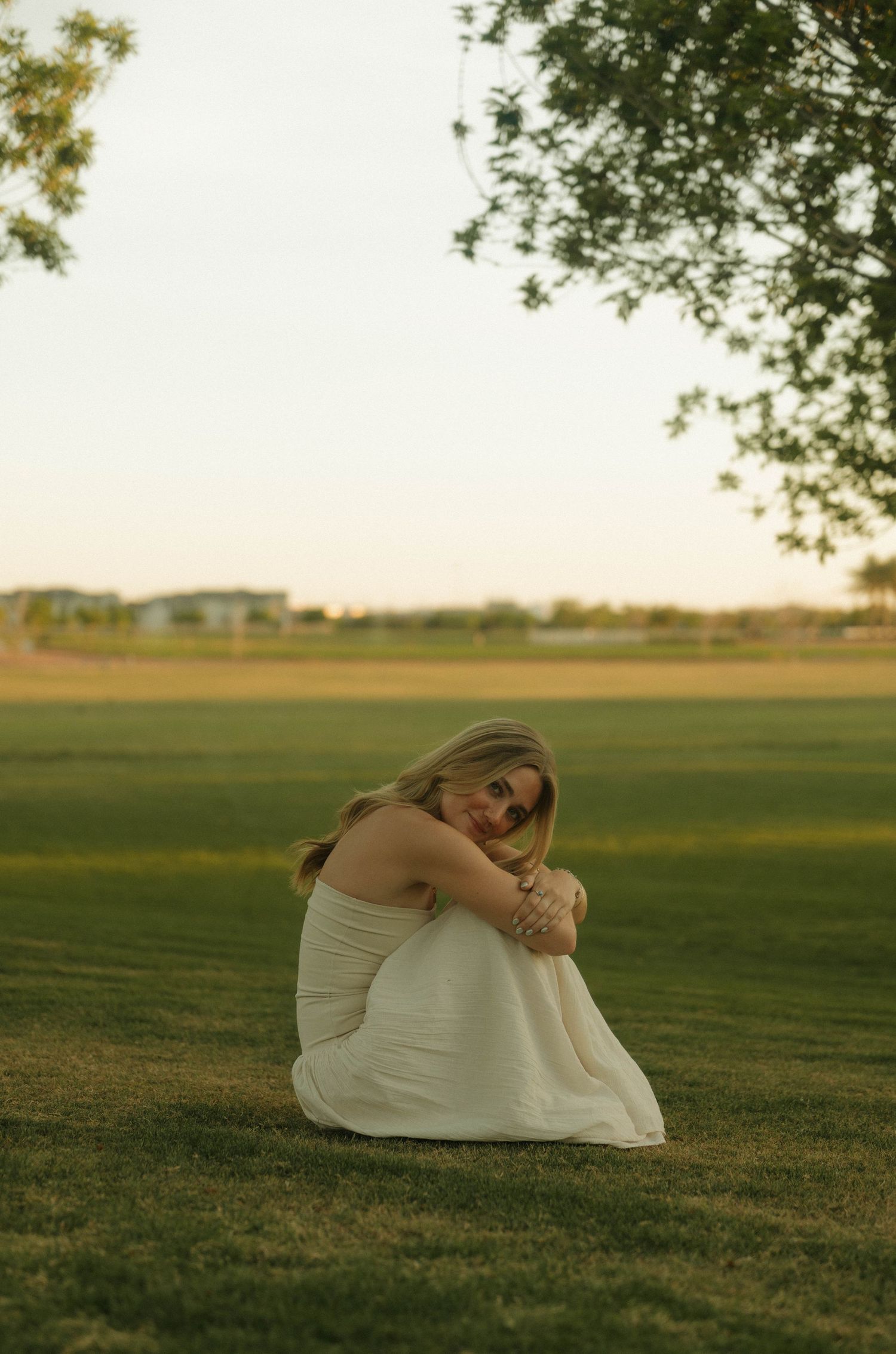 Blonde woman in white dress sitting on green grass hugging her knees at golden hour.