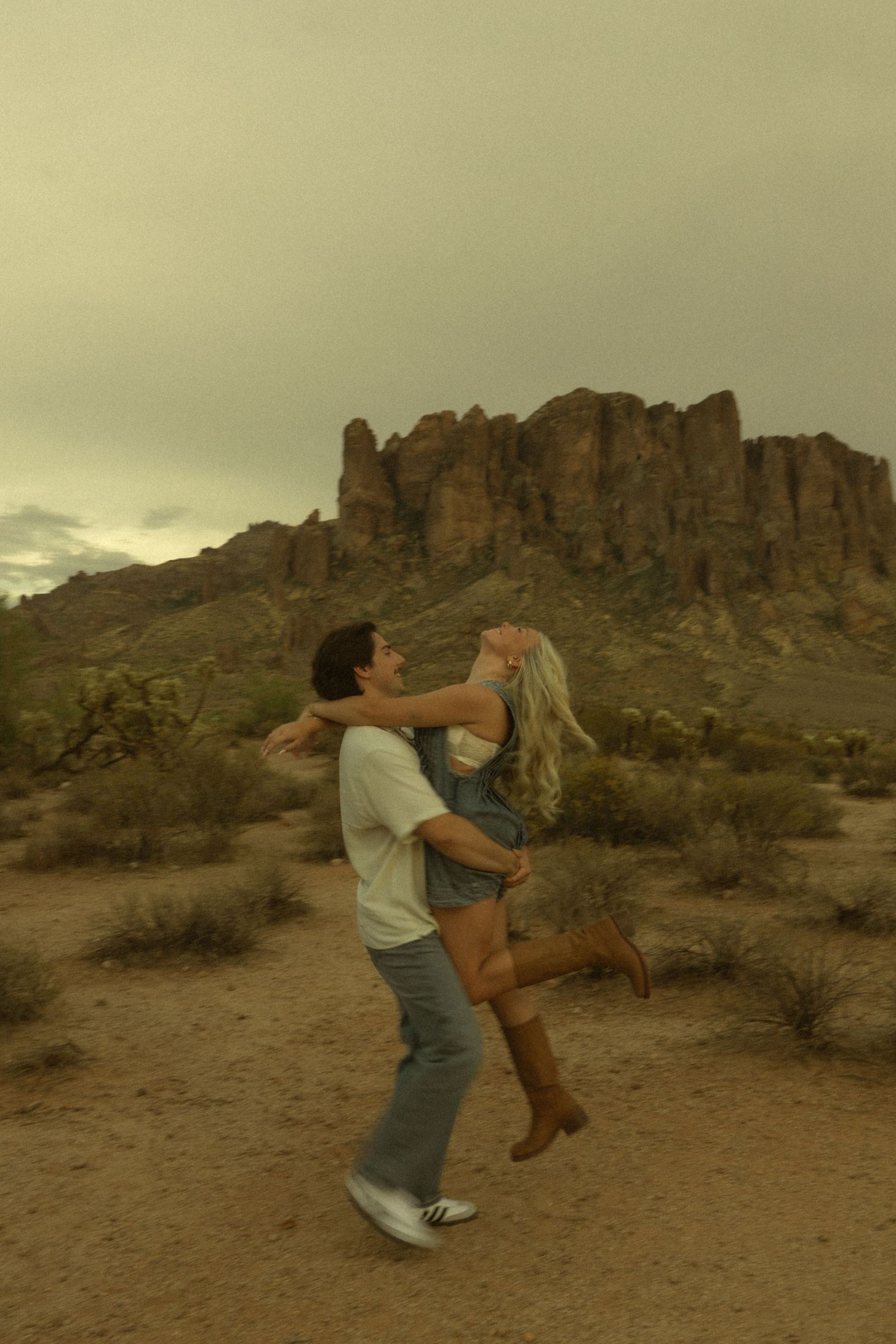 Couple embracing and spinning in desert landscape with rocky mountains and cacti under cloudy sky.