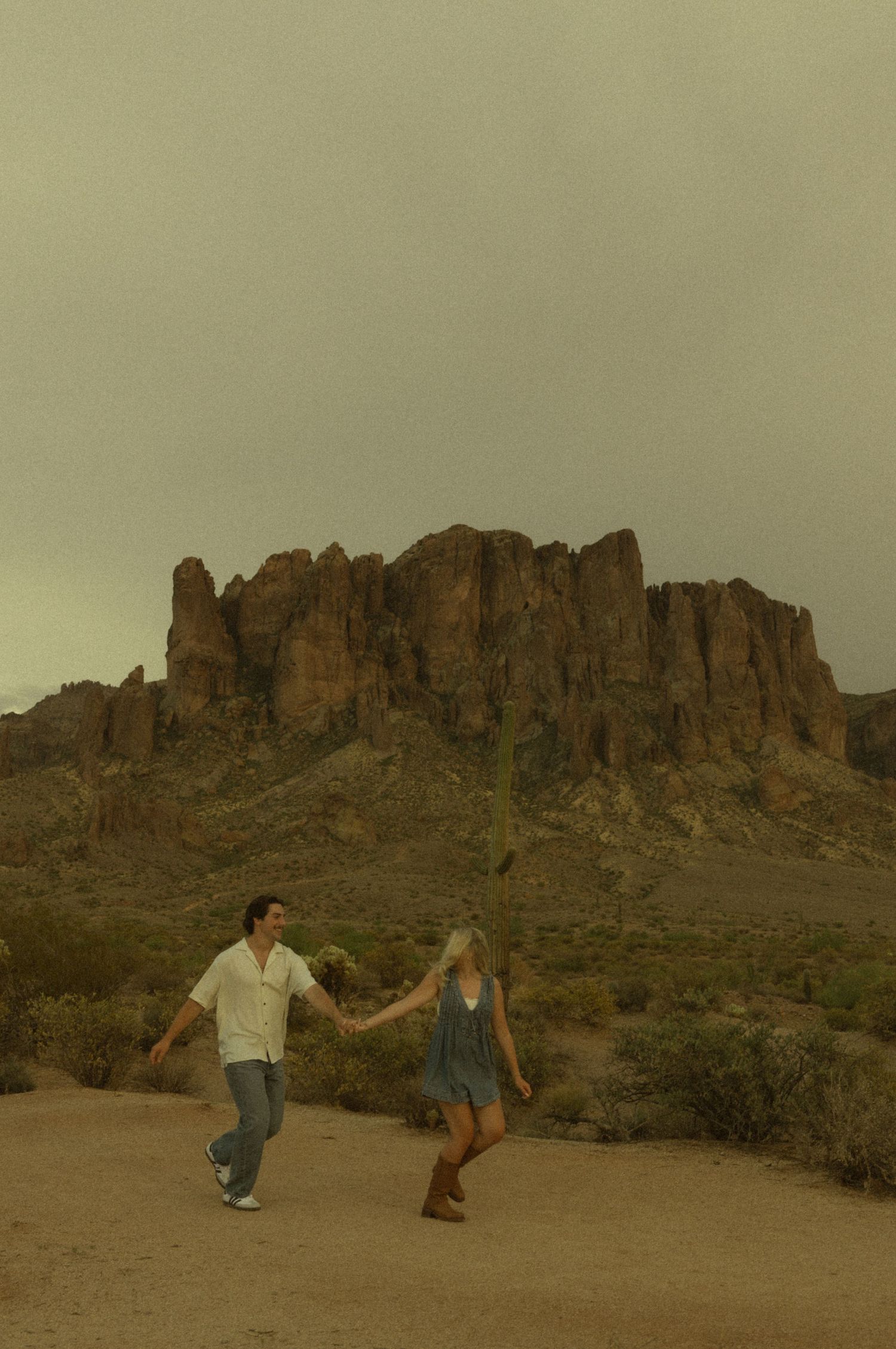 Couple running hand in hand on desert trail with Superstition Mountains in background.