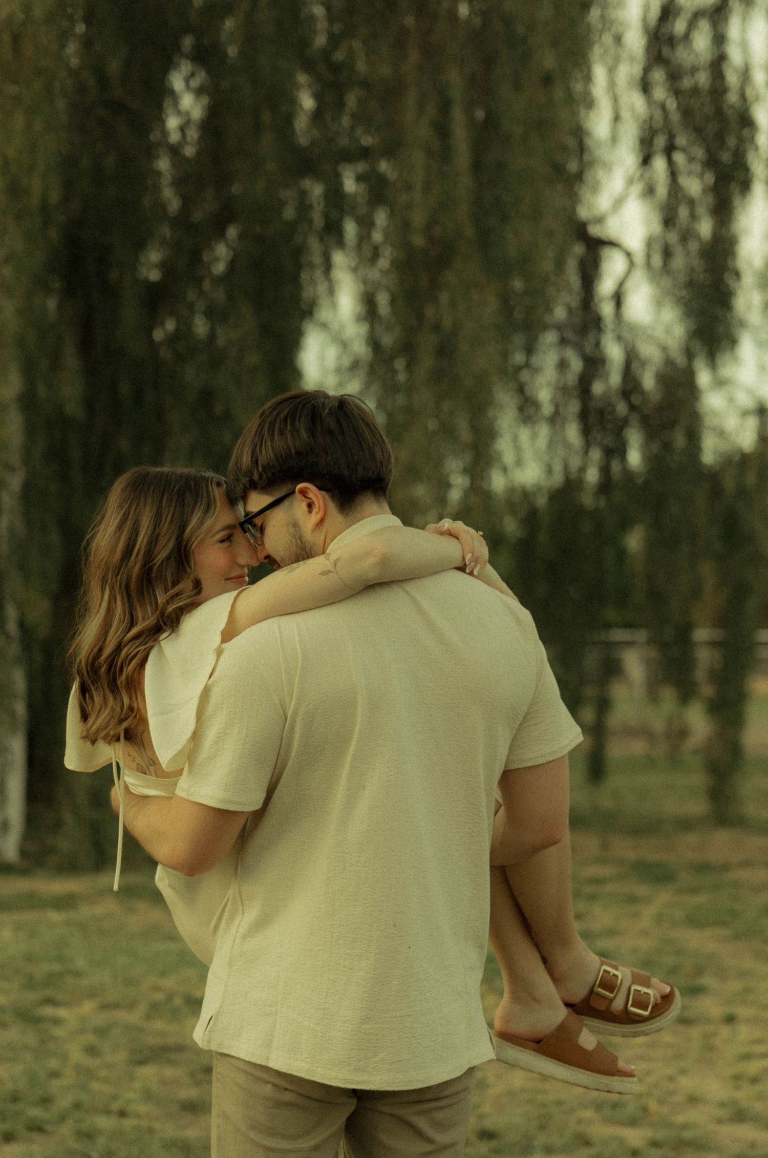 A couple embraces romantically outdoors near a weeping willow tree, the man lifting the woman in a tender hug.