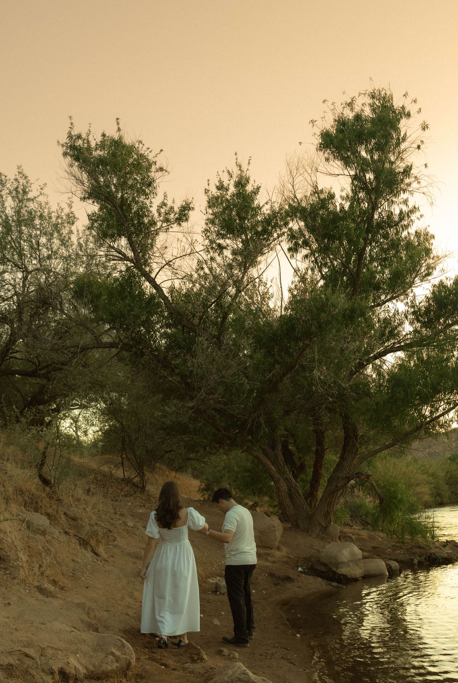 Couple holding hands walking along sandy riverbank at dusk near green trees.