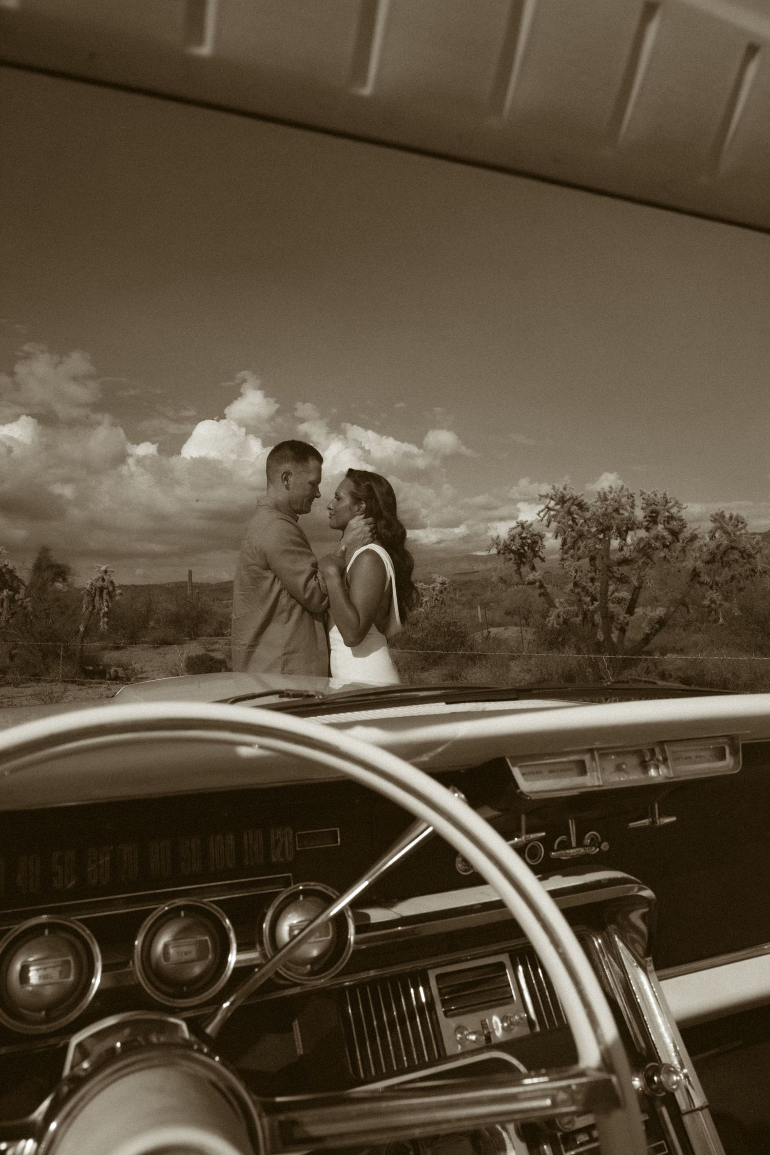 Sepia-toned photo of a couple embracing outdoors, framed through a vintage car's windshield and steering wheel.
