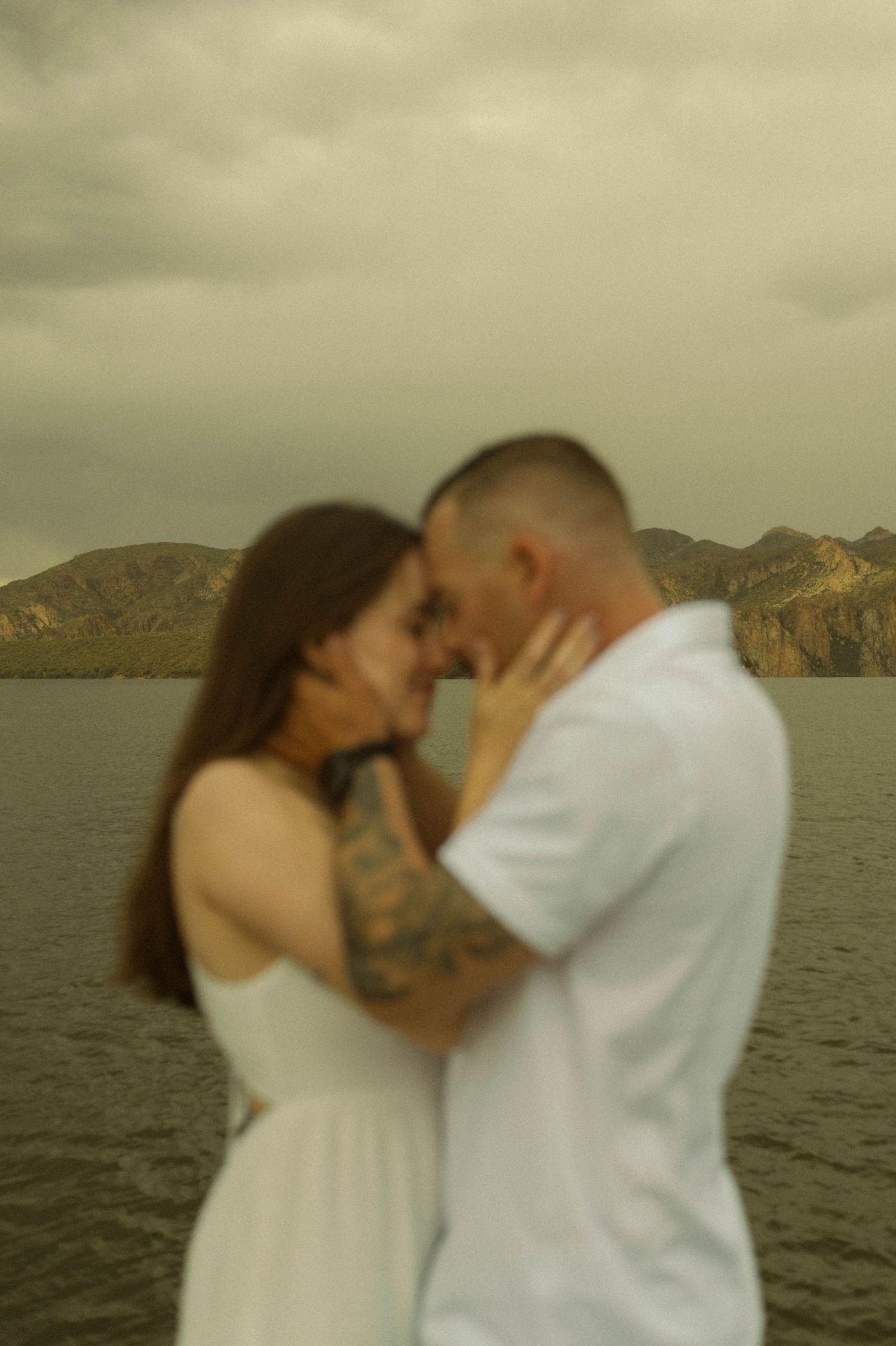 An intimate embrace between a couple against a moody lakeside mountain landscape.