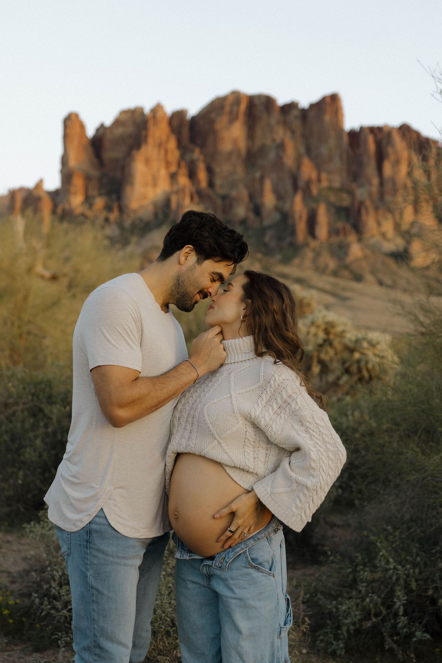 An expecting couple shares an intimate moment against a backdrop of desert mountains.