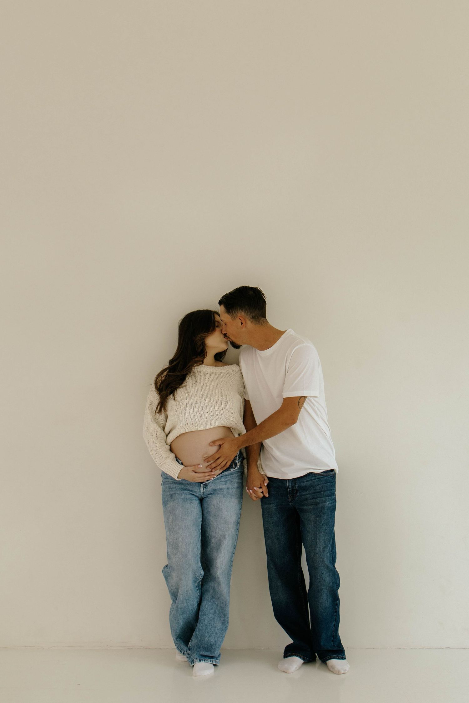 A couple in casual attire shares an intimate moment against a beige wall, highlighting her pregnant belly.