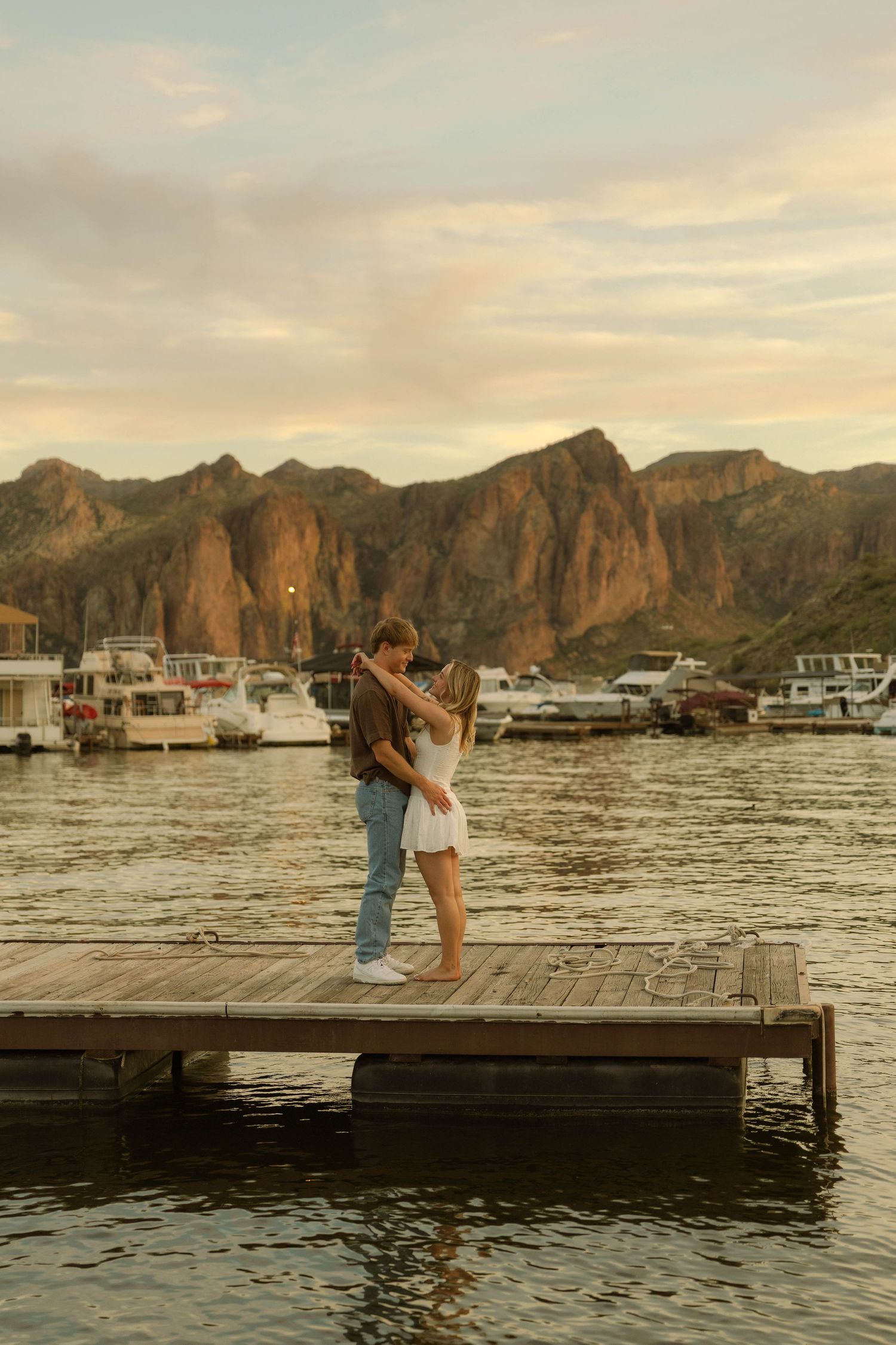 A couple embraces on a wooden dock with boats and dramatic mountain scenery at golden hour.