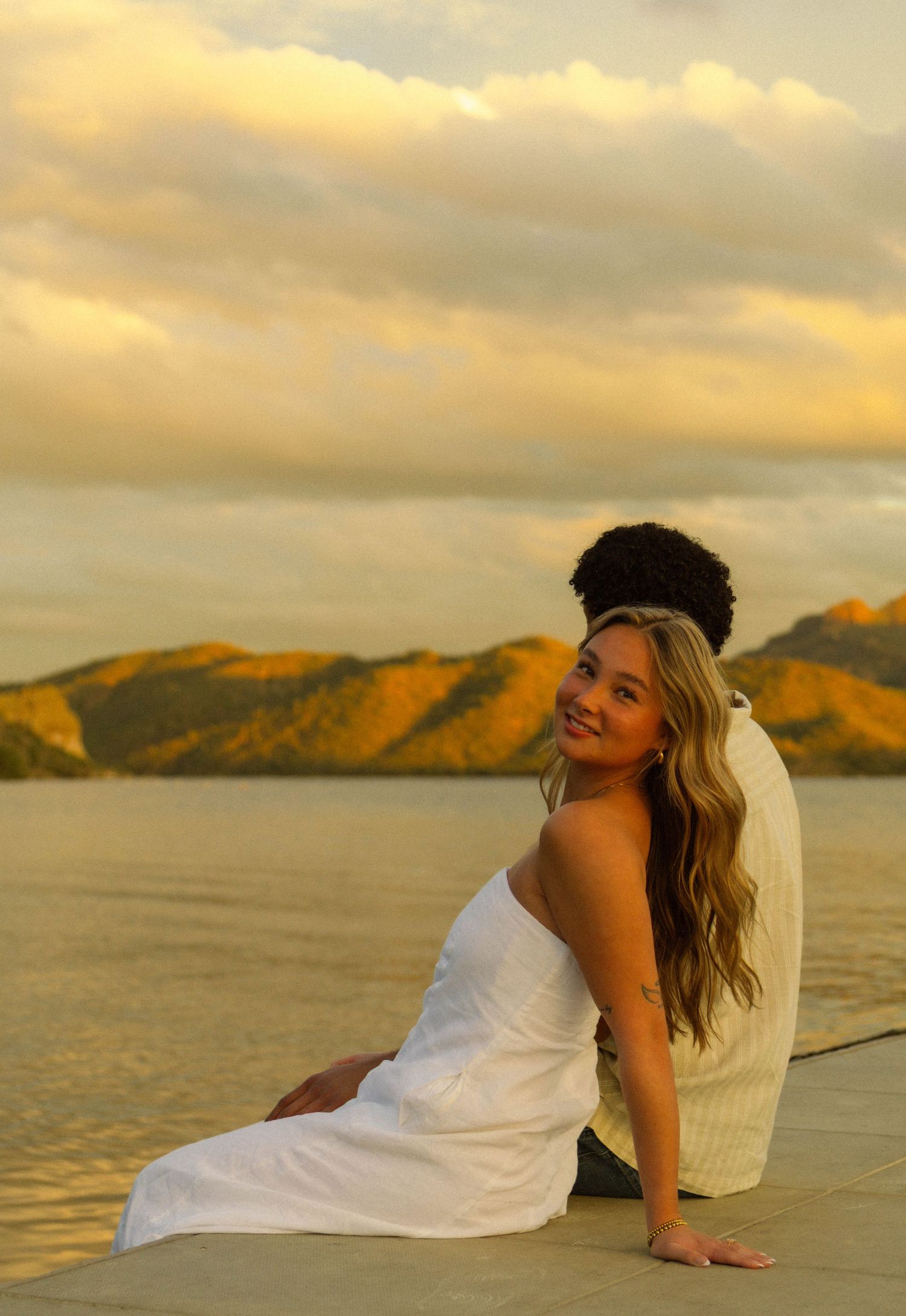 A person in a white dress poses gracefully on a beach at sunset with mountains and ocean in the background.