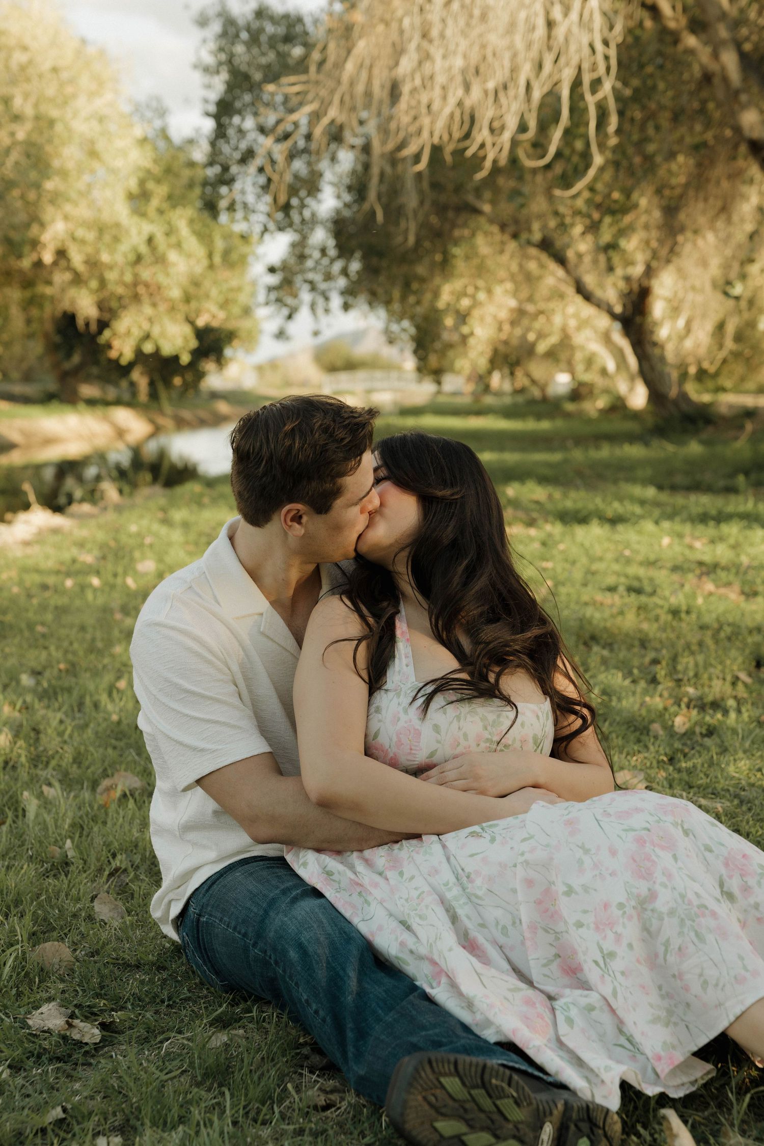 A romantic couple shares tender kisses while laying in a sunlit grassy field on a warm afternoon.